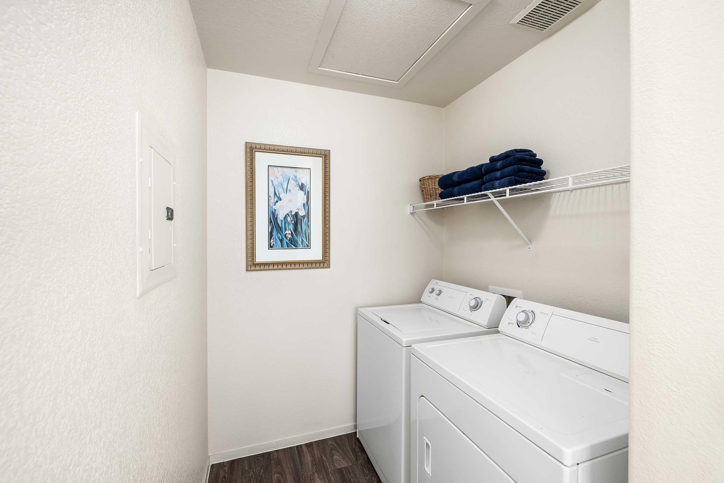 A laundry room featuring two white washing machines side by side, a framed piece of artwork on the wall, and neatly folded blue towels on a shelf above. The room has light-colored walls and dark wood-like flooring, creating a clean and organized space for laundry tasks.