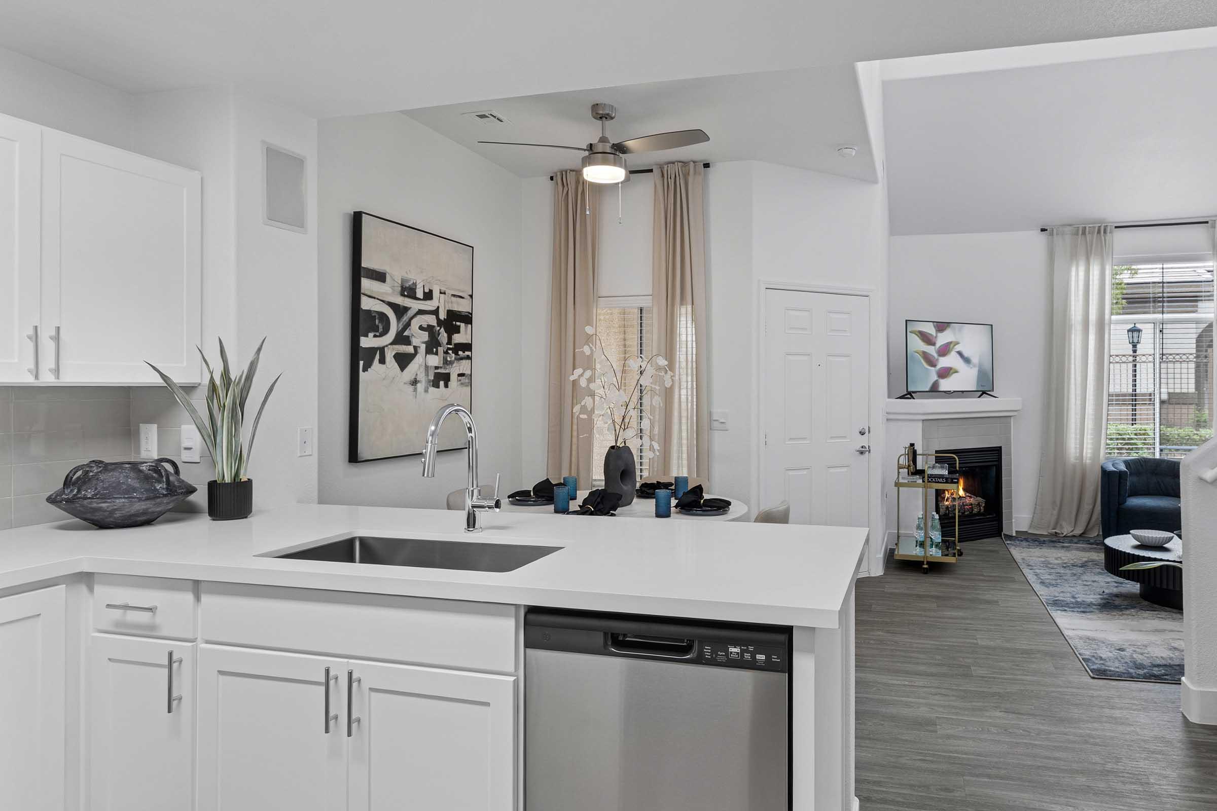A modern kitchen with white cabinets and a stainless steel dishwasher. In the background, a comfortable living area features a blue sofa, decorative plants, and wall art. Large windows with sheer curtains allow natural light to fill the space.
