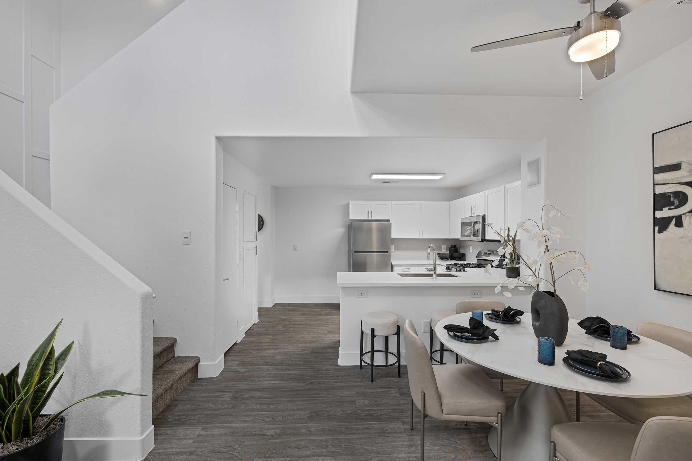 A modern kitchen and dining area featuring a round table set with black plates and blue glasses. White cabinetry and appliances are visible in the kitchen, along with a plant in the corner. A staircase leads to an upper level, and there’s a large piece of abstract art on the wall. Bright and minimalist decor.