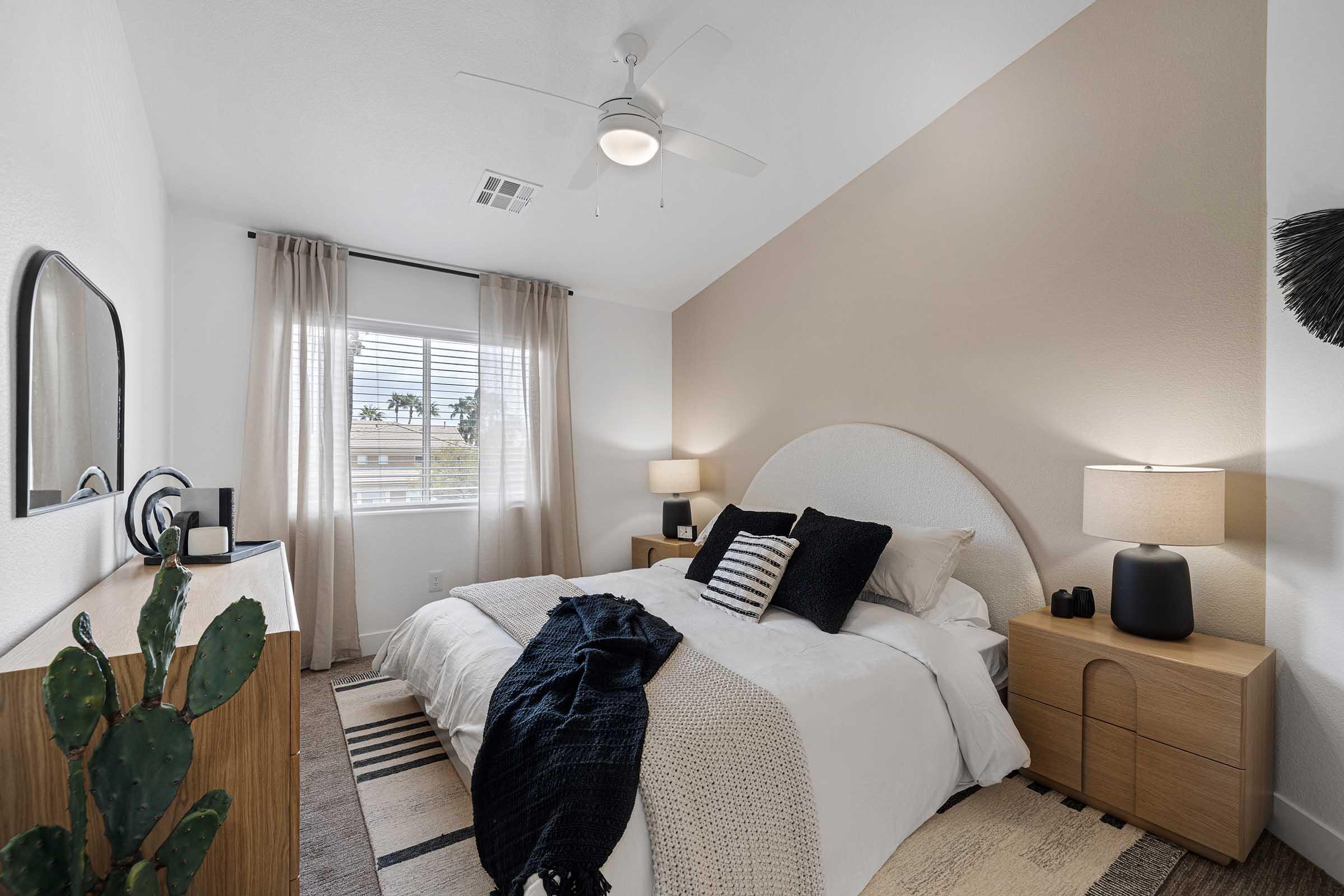 A cozy bedroom featuring a queen-sized bed with white bedding and decorative pillows. There are two lamps on bedside tables, a wall-mounted mirror, and a potted cactus near a window with sheer curtains. The walls are painted in soft, neutral tones, and a ceiling fan is installed.