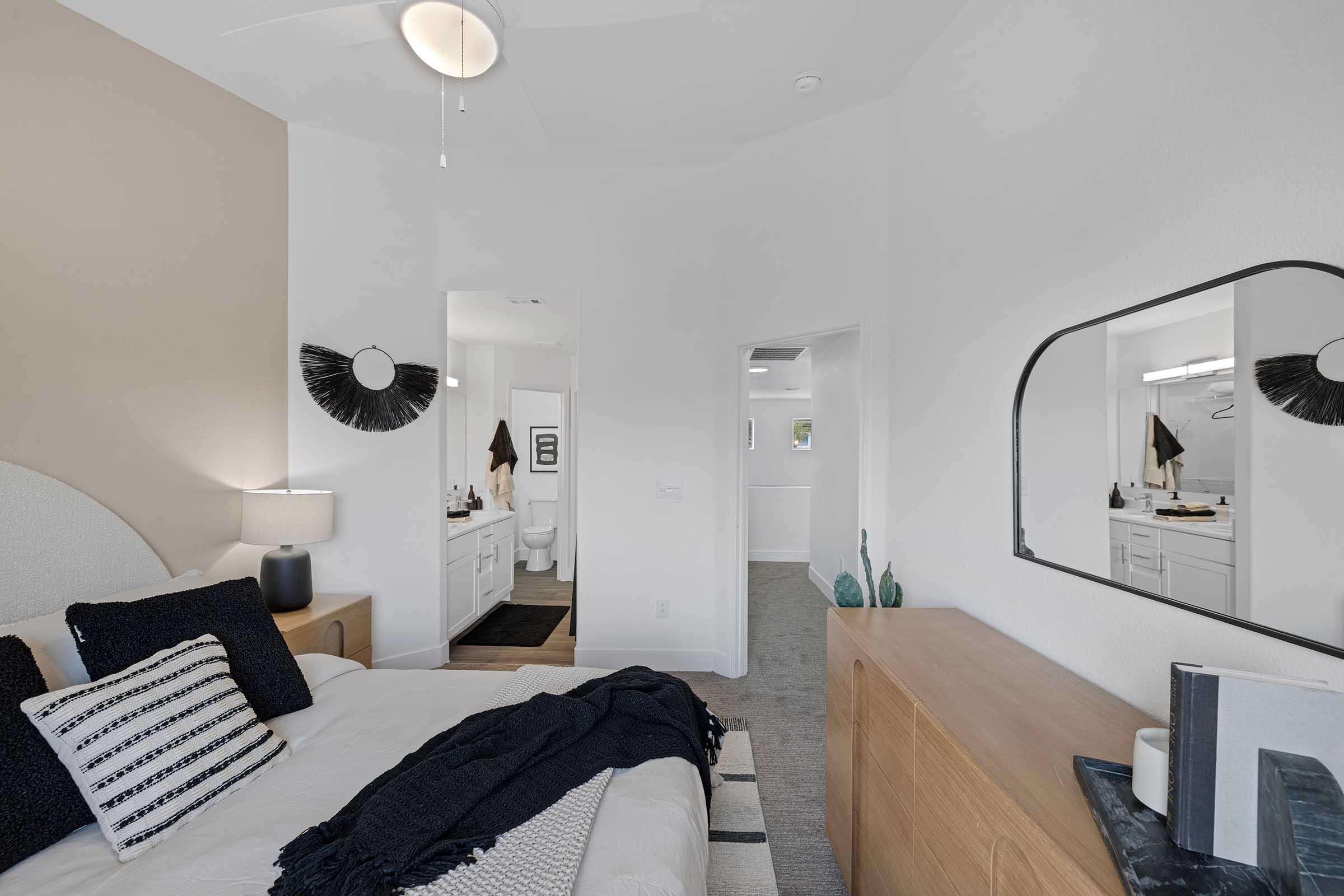 A modern bedroom featuring a queen-sized bed with dark bedding, a stylish light fixture, and a wooden dresser. The room has a warm color palette with neutral walls and a decorative mirror. A doorway leads to an ensuite bathroom, providing a spacious and inviting atmosphere.
