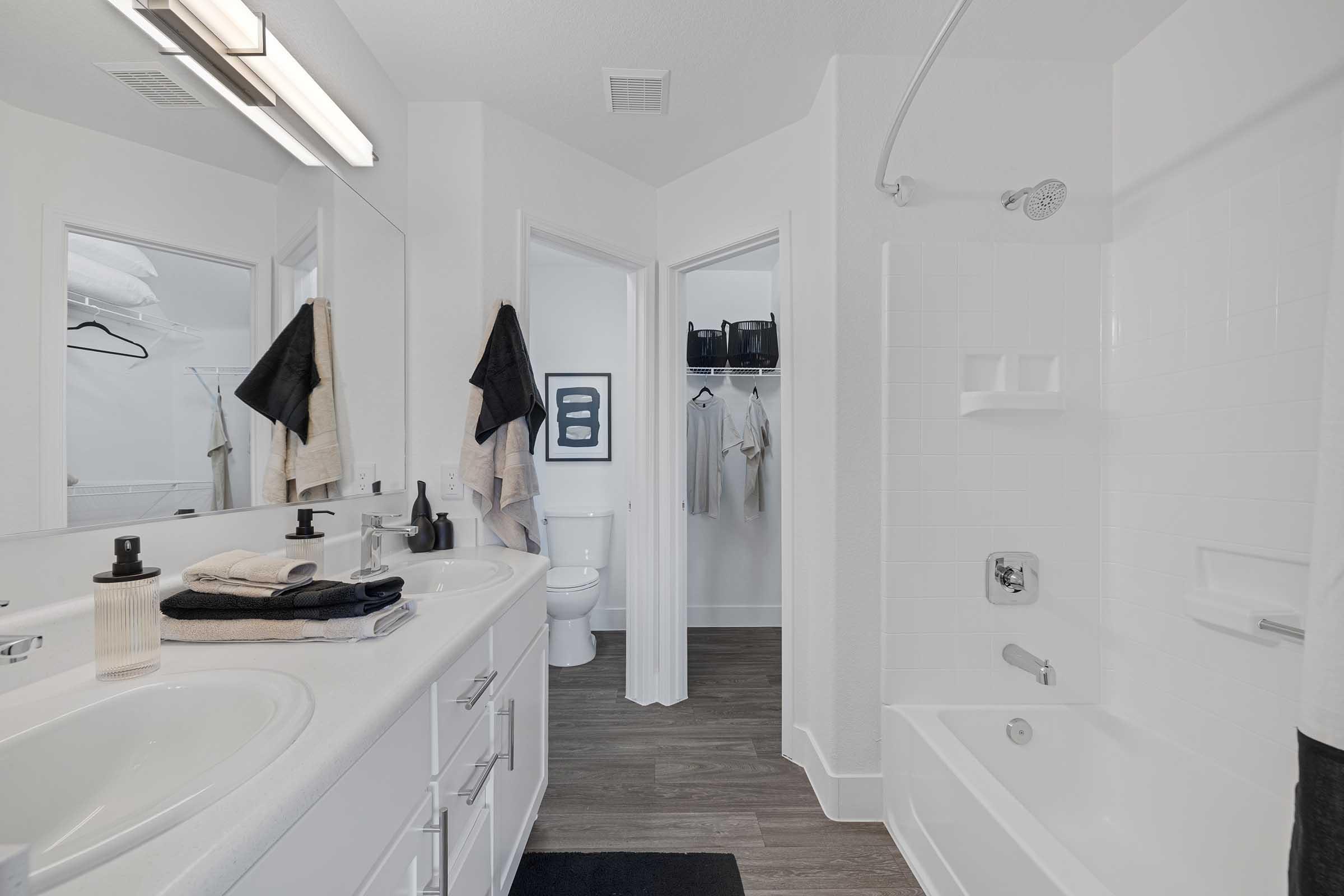 A modern bathroom featuring a double vanity with sinks, white cabinetry, and neatly arranged towels. The space includes a bathtub with a shower, a toilet in a separate area, and a walk-in closet visible in the background. The decor is minimalistic with a neutral color palette and wooden flooring.