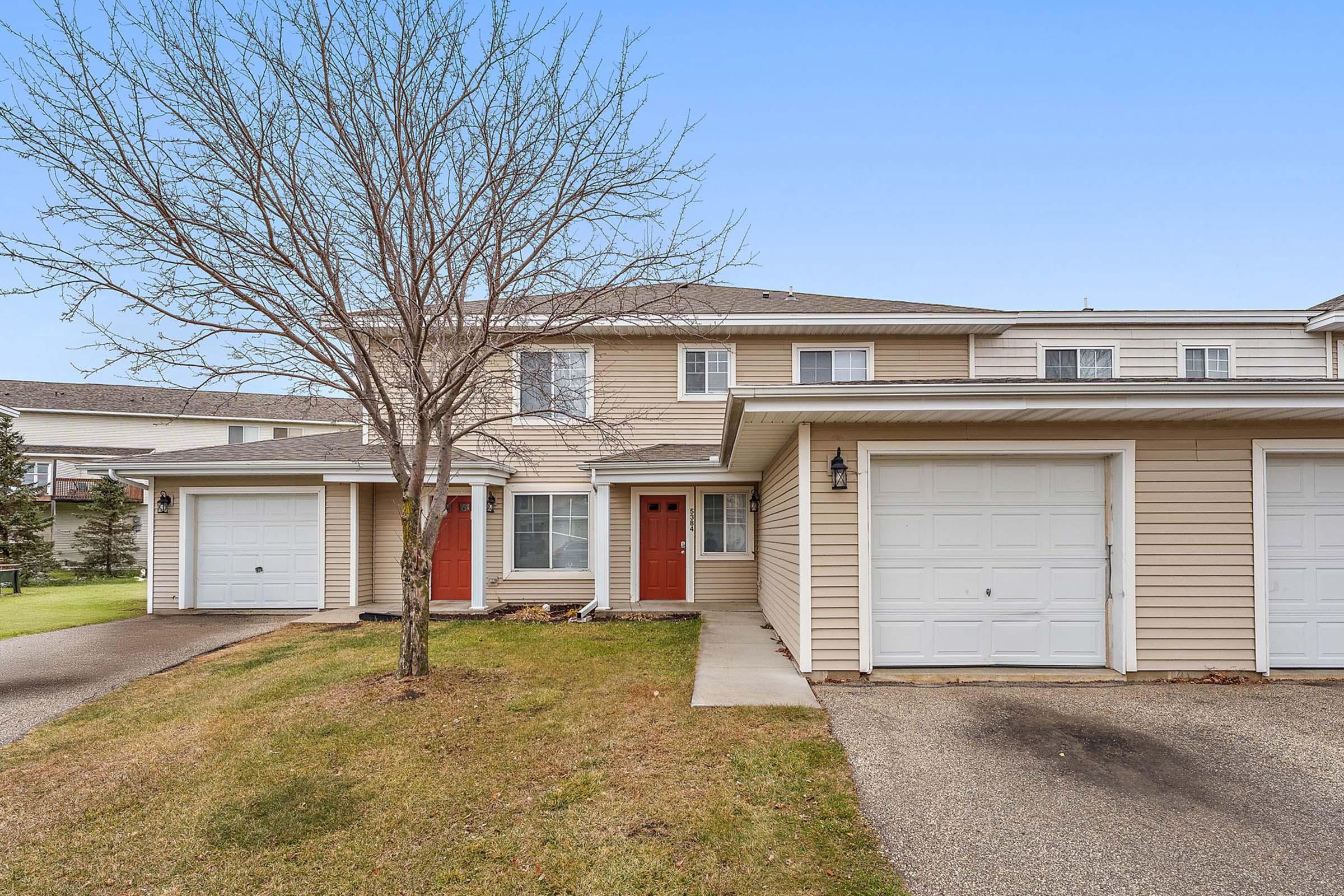 A two-story residential building with beige siding and red doors. It features two garage doors on the left side, a small front yard with a tree that is bare of leaves, and a walkway leading to the entry. The sky is clear and blue, indicating a pleasant day.