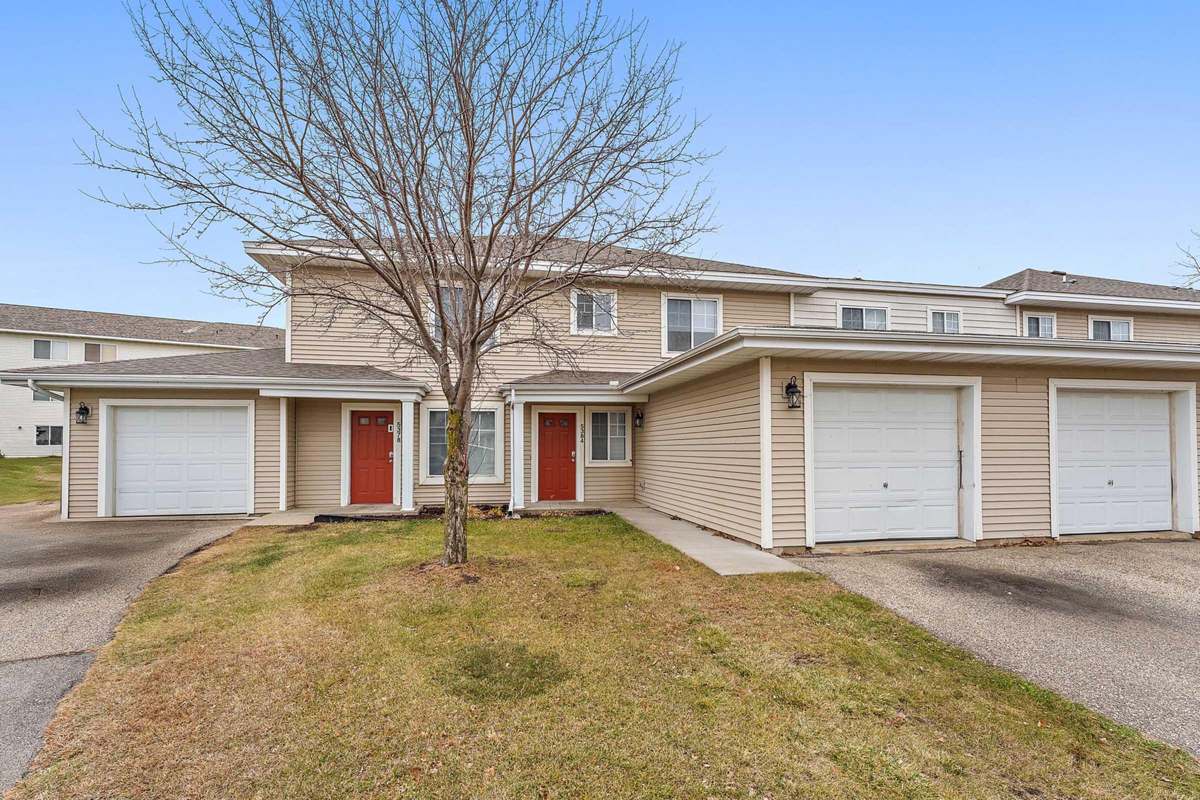 A two-story residential building featuring beige siding, two front doors painted in red, and attached garages. A bare tree stands in the front yard, with grass and a paved walkway visible. The background includes additional residential structures under a clear blue sky.