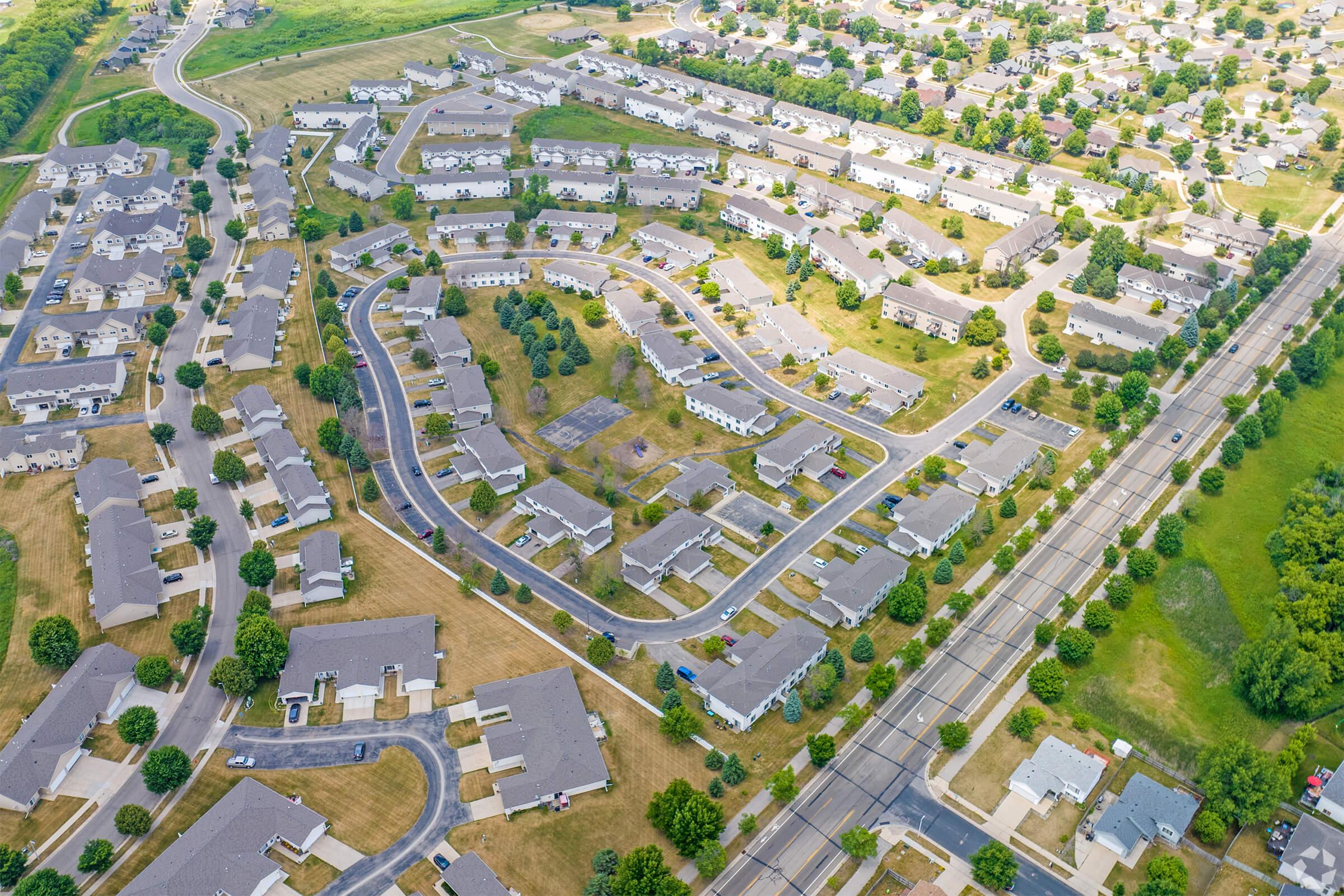 Aerial view of a suburban neighborhood featuring a grid of residential homes, green lawns, and tree-lined streets. Some areas are under construction or vacant, while others are fully developed. A road runs along the edge, connecting to the community. The landscape includes patches of grass and trees, highlighting the organized layout of the area.