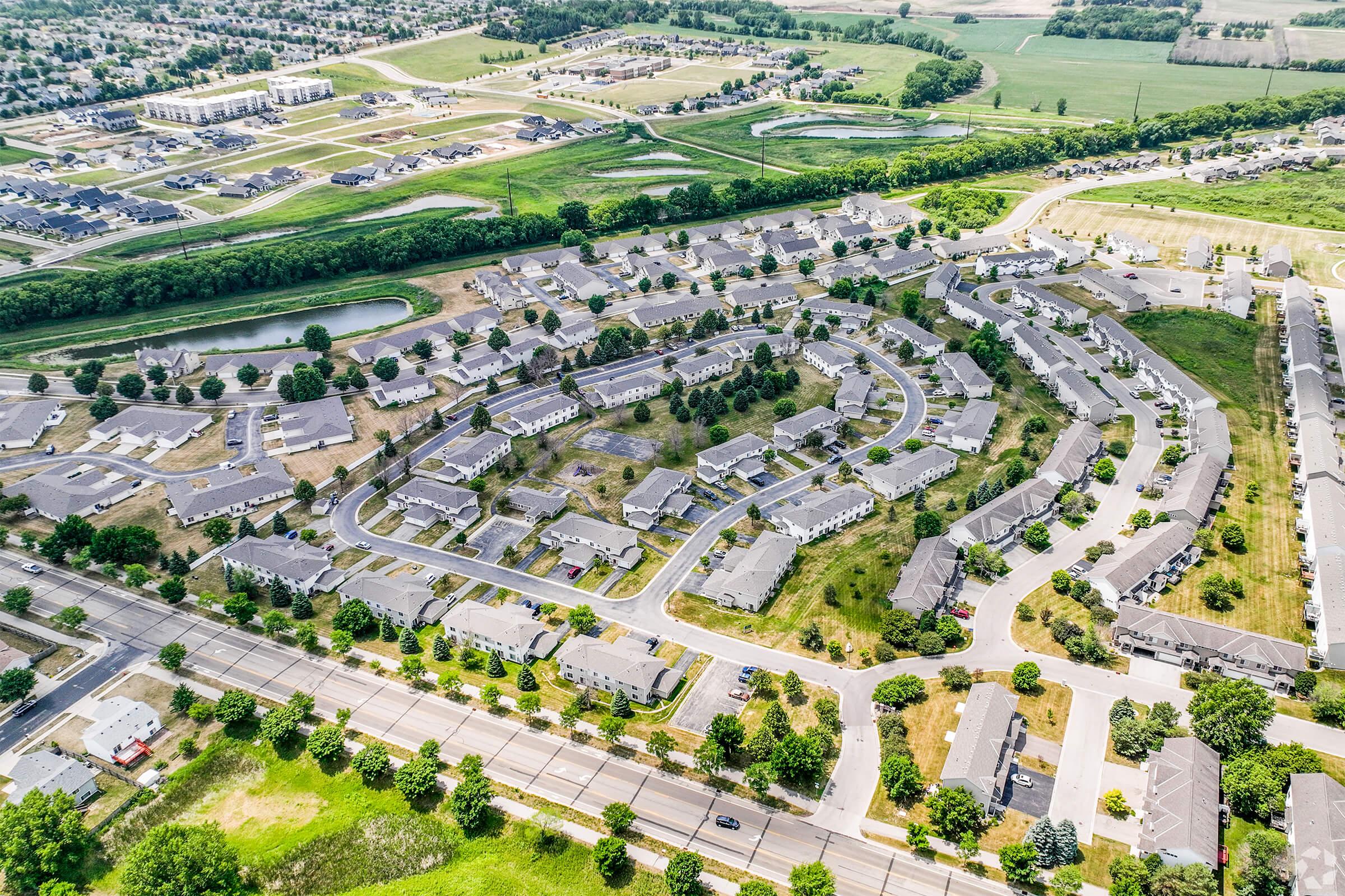 Aerial view of a suburban neighborhood featuring curved streets and clusters of residential buildings. Green spaces and a small pond are visible, along with nearby commercial areas and open fields in the background. The layout reflects organized housing development in a rural setting.