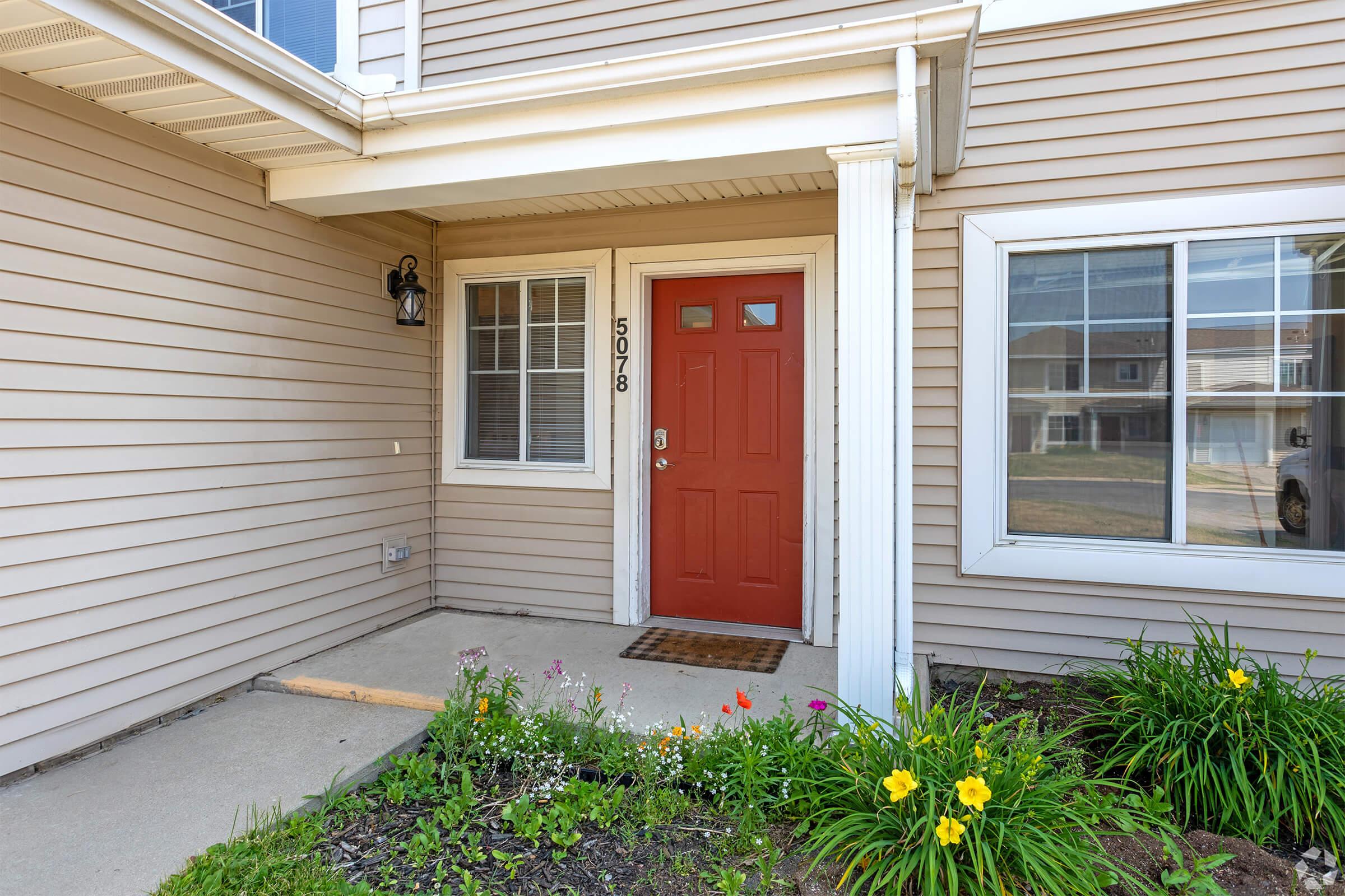 A welcoming front entrance featuring a bright red door with the number 5078 displayed, flanked by two windows. The porch is decorated with blooming flowers and green foliage, creating a vibrant and inviting atmosphere. The home's exterior is a soft beige siding, adding to the charm.