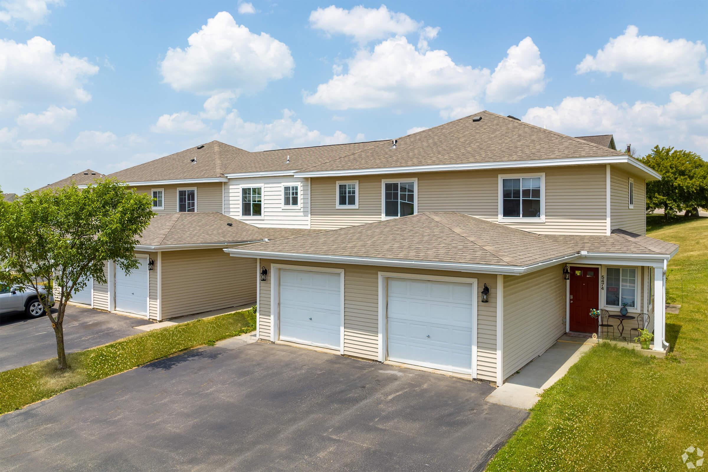 A two-story residential building with beige siding and multiple garages. The property features a well-manicured lawn and is situated under a blue sky with scattered clouds. Parking spaces are visible in front of the garages.