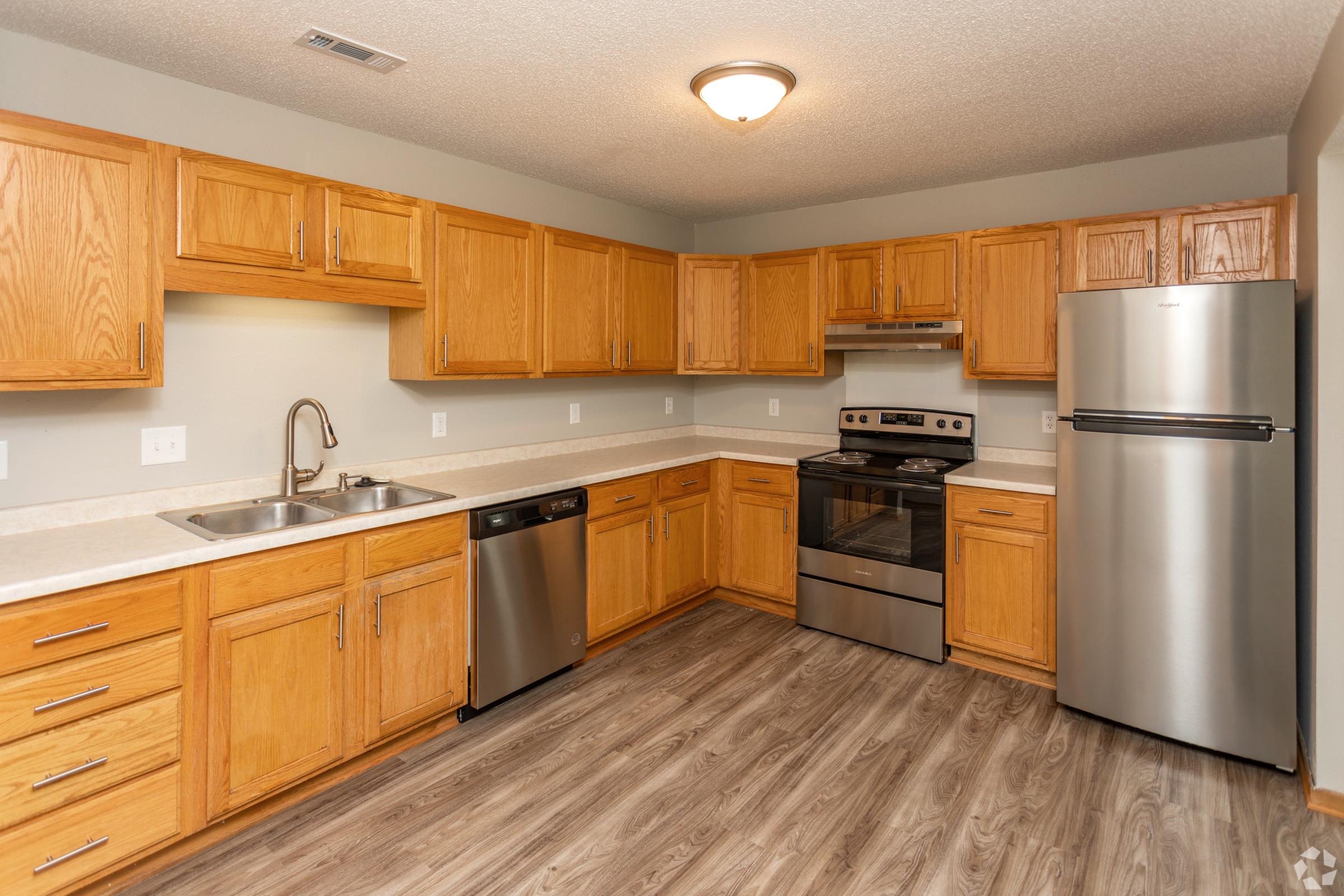 Modern kitchen featuring wooden cabinets, stainless steel appliances including an oven, microwave, and refrigerator. The countertop is light-colored, and there is a double sink with a faucet. The floor is a wood-like laminate, and the walls are painted in a neutral tone, creating a bright and inviting atmosphere.