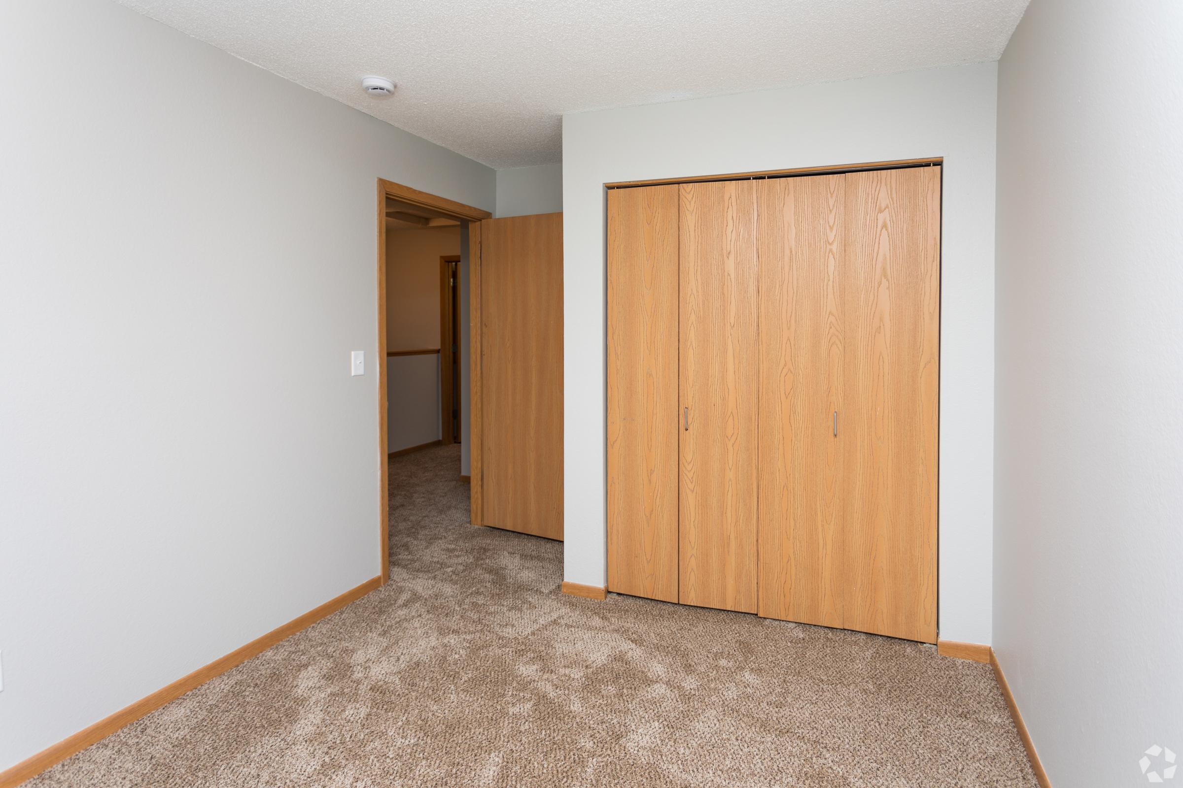 Interior shot of a room featuring beige carpet flooring, light gray walls, and a pair of open wooden closet doors. The space is well-lit and appears empty, with a door to another area visible in the background.