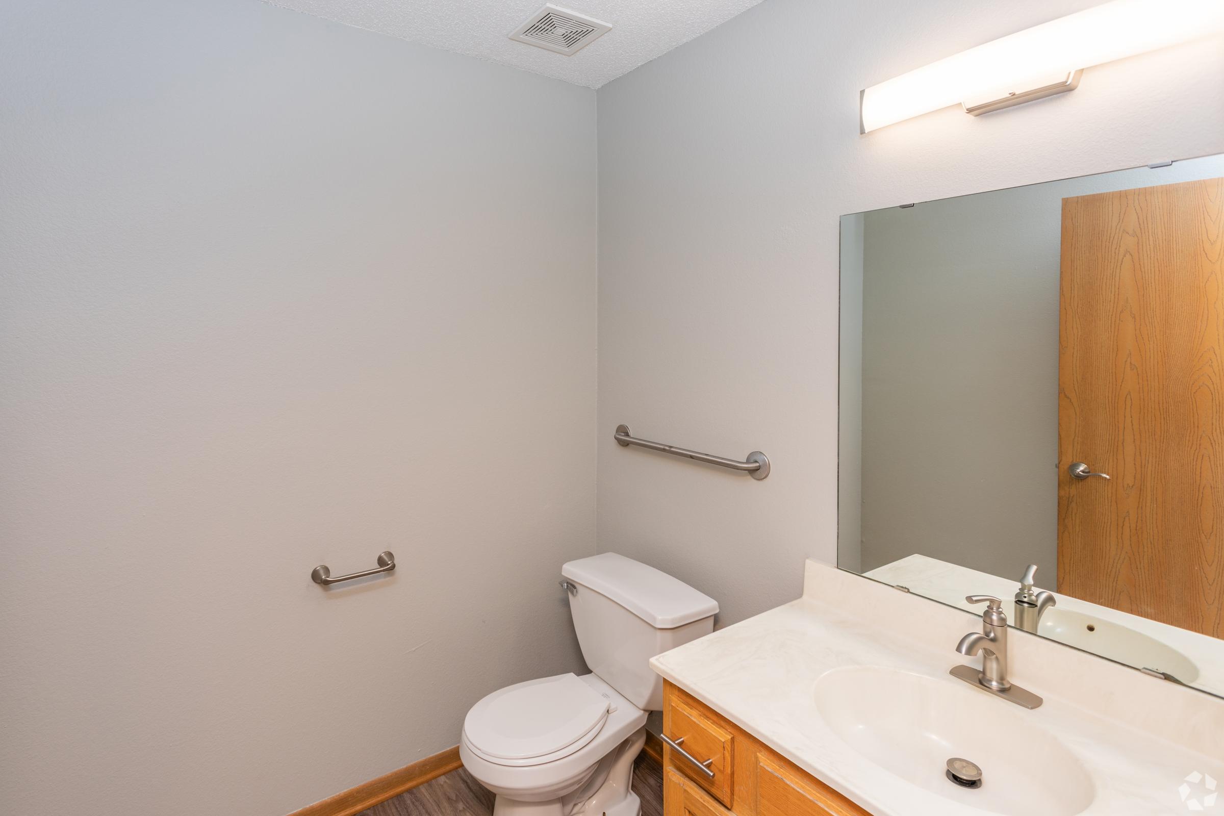 A clean, simple bathroom featuring a white toilet, a single sink with a faucet, and a large mirror above the sink. The walls are painted in a light gray, and there is a wooden cabinet beneath the sink. A towel rack is mounted on the wall, providing accessibility. The floor is finished with light-colored vinyl.