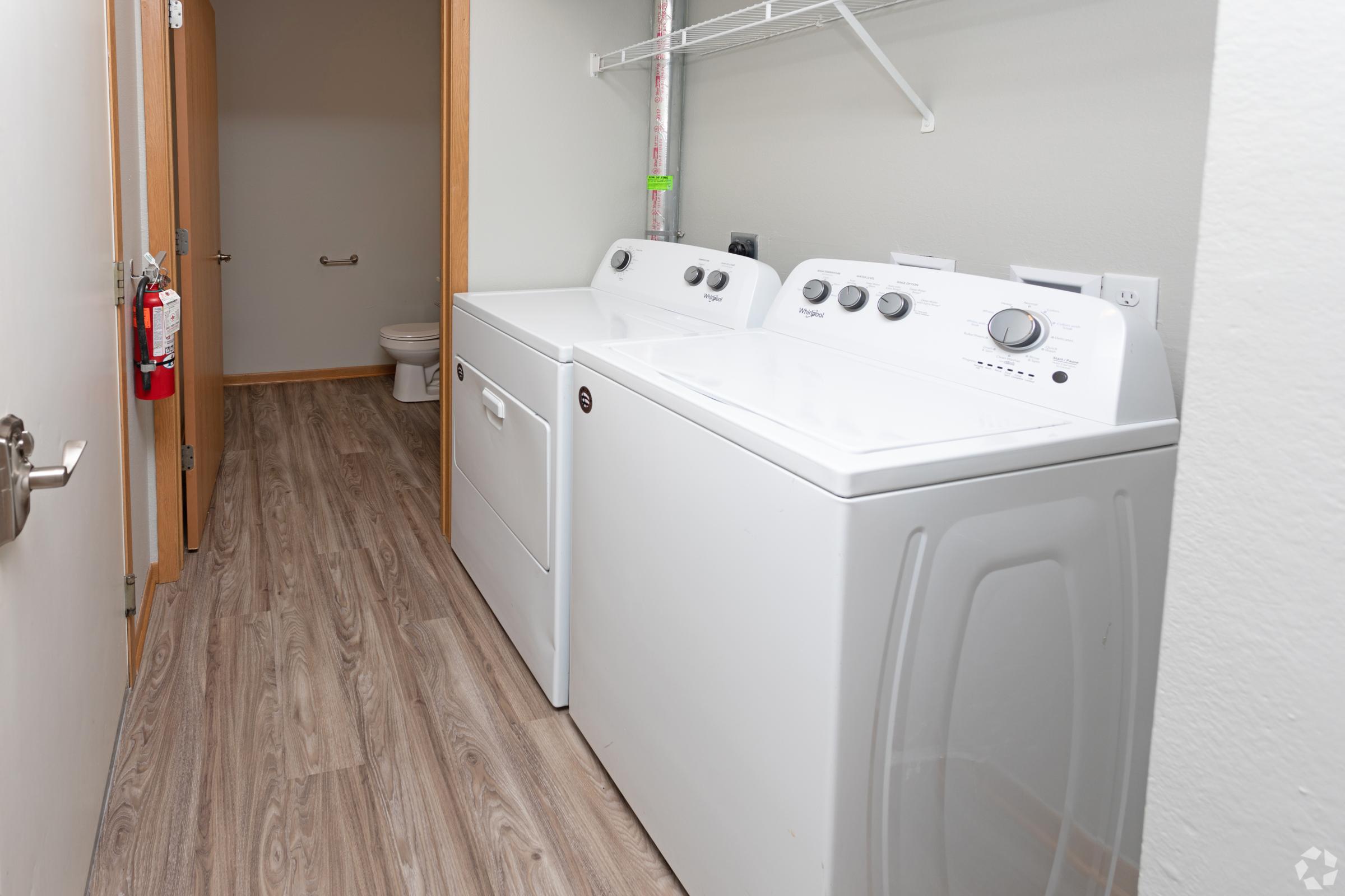 A laundry room featuring a white washing machine and dryer side by side. The room has light-colored walls and a wooden floor. There is a shelf above the appliances for storage. In the background, a door leads to a bathroom with a toilet visible. A fire extinguisher is mounted on the wall.