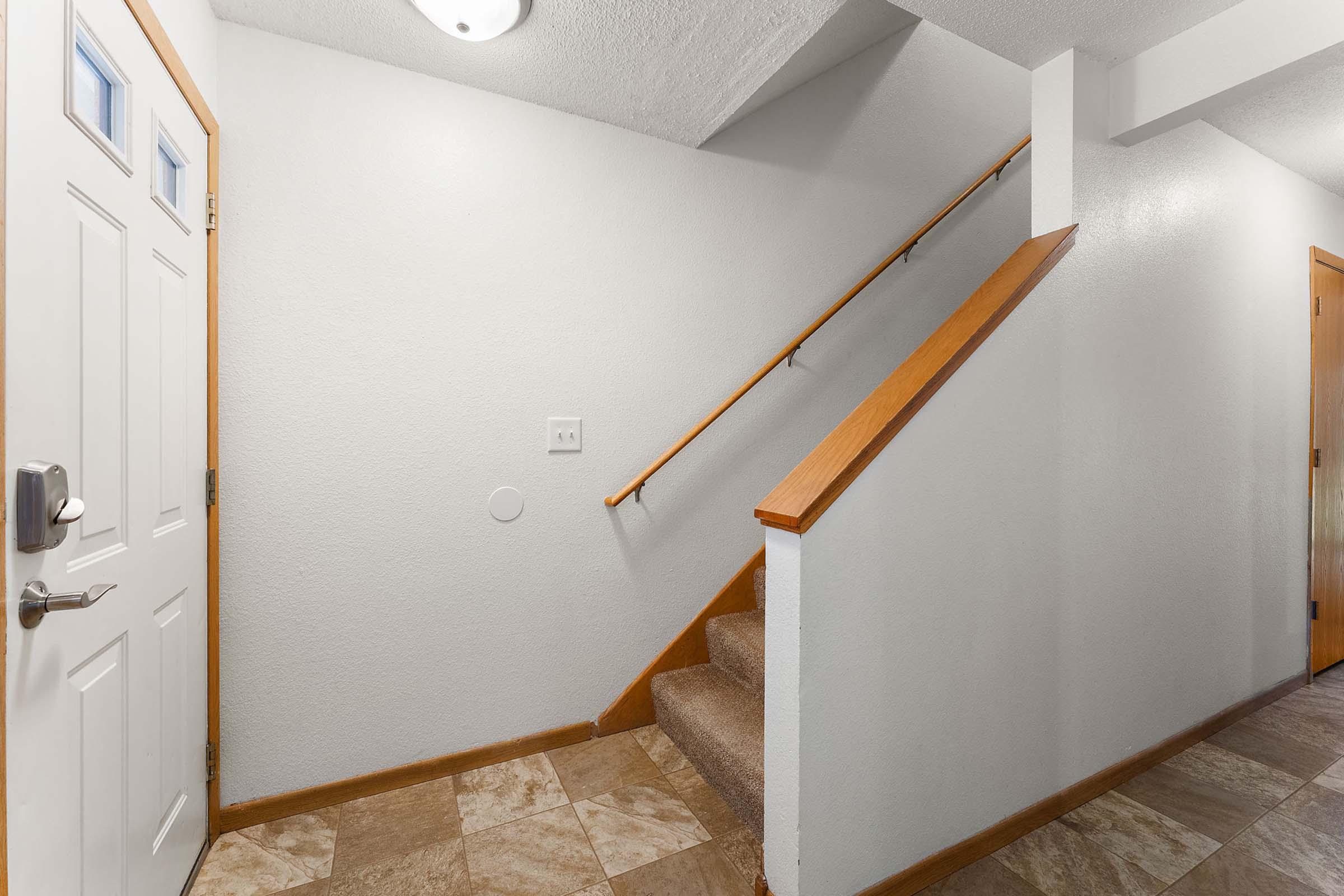 A well-lit entryway featuring a white door, light-colored walls, and a staircase with wooden handrails. The floor is tiled with a large pattern, and there is a small circular wall feature near the base of the stairs. The space is clutter-free and offers a welcoming atmosphere.