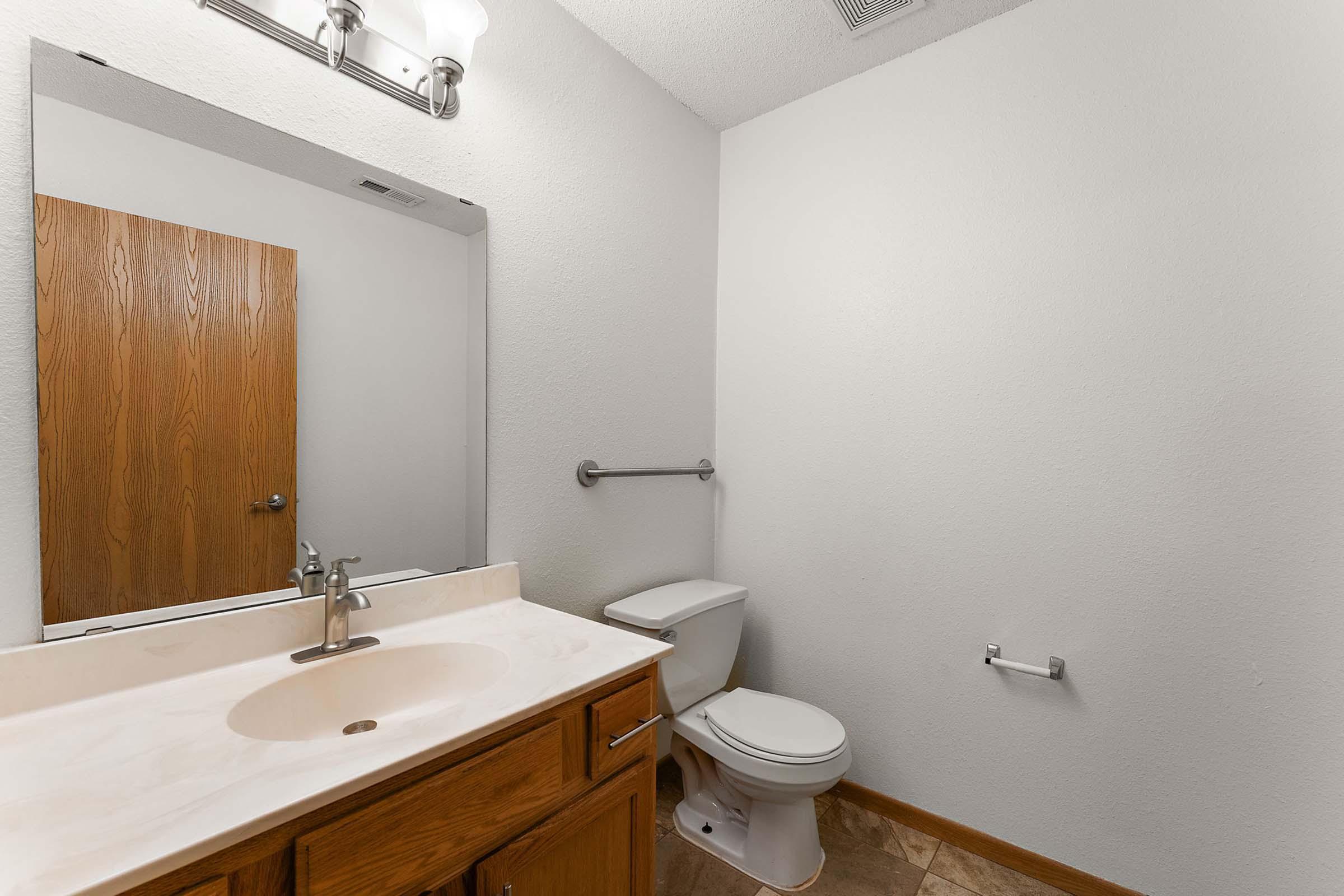 A small, modern bathroom featuring a light-colored countertop with a sink, a toilet, and a single grab bar on the wall. A large mirror reflects the space, and there's a wooden door visible in the background. The walls are painted white, and the floor has light-colored tiles.