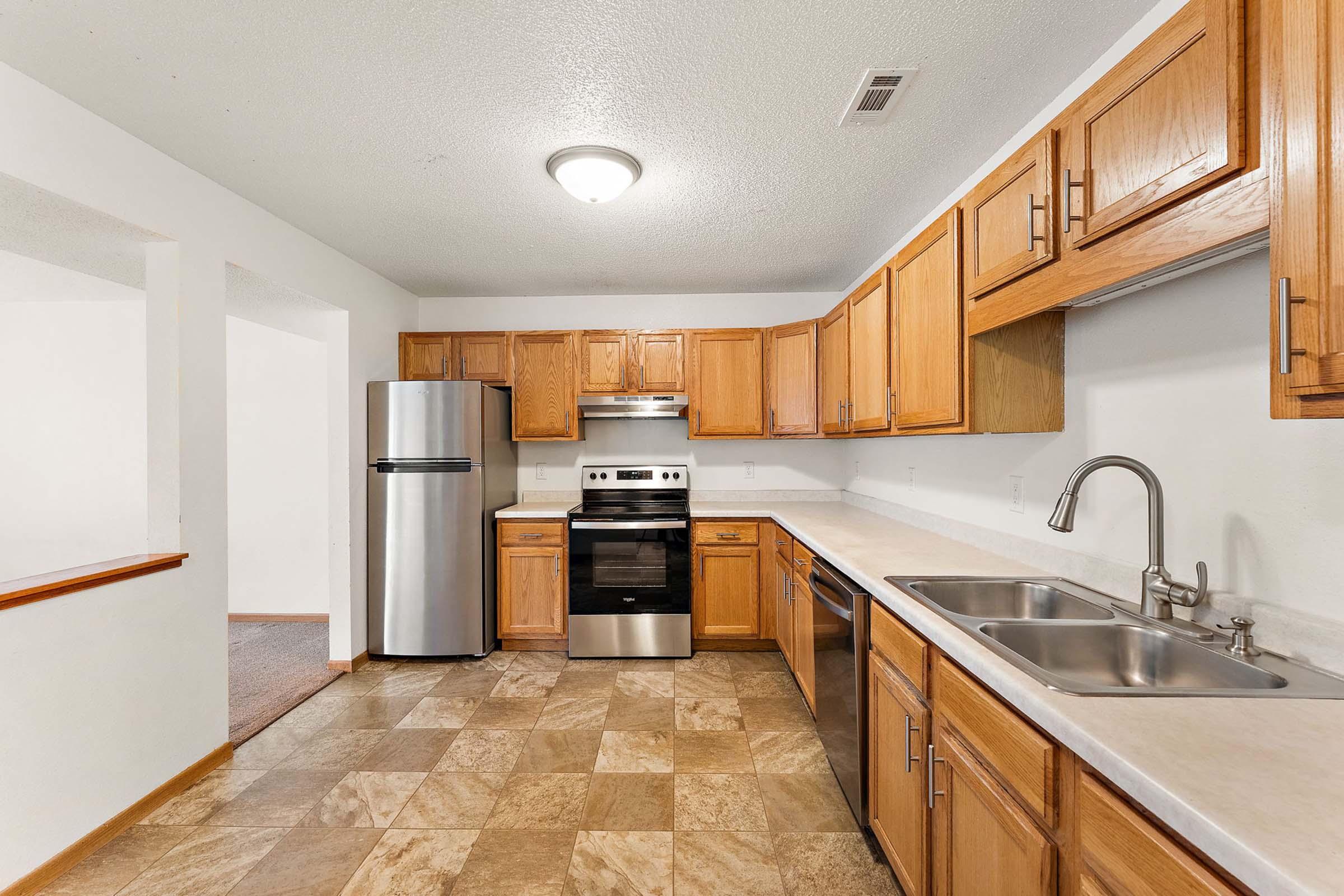 A spacious kitchen featuring wooden cabinets, stainless steel appliances, and a double sink. The floor is covered with patterned tiles, and there is a refrigerator to the left. The kitchen is well-lit with a ceiling light, and the layout includes an open space leading to another room.