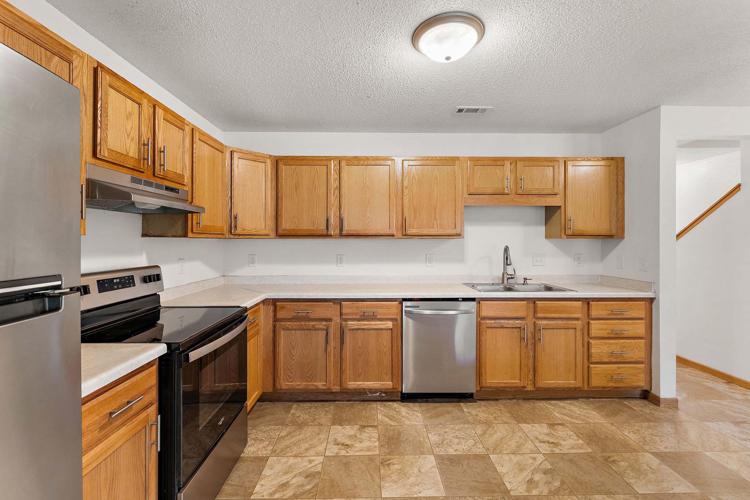 A modern kitchen featuring light wood cabinets, stainless steel appliances including a refrigerator, oven, and dishwasher, and a spacious countertop. The floor is tiled with square, light-colored tiles, and there is a single bowl sink under a window. The room is well-lit with natural light.