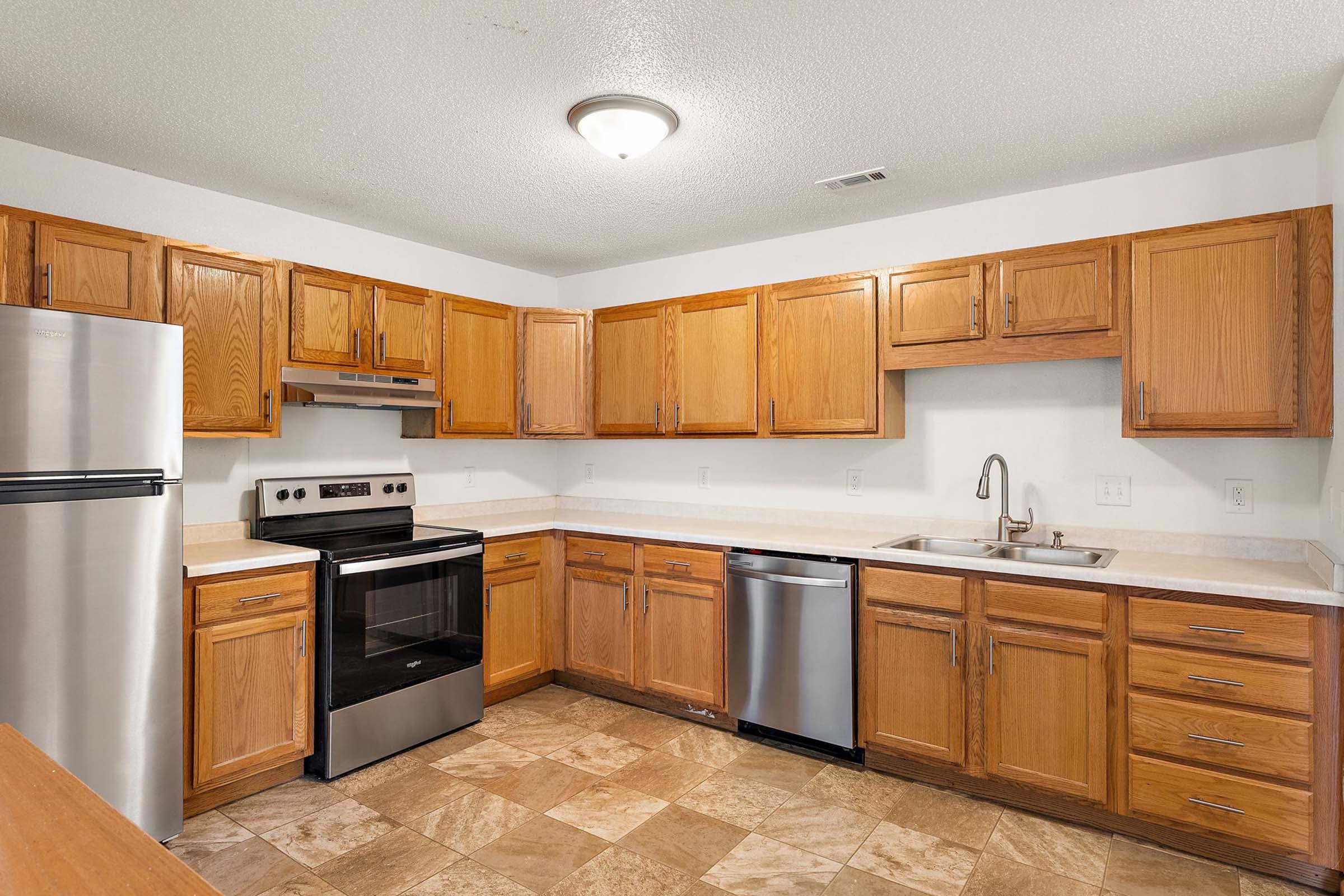 A modern kitchen featuring wooden cabinetry, stainless steel appliances including a refrigerator, stove, and dishwasher. The countertop is light-colored, and the flooring consists of beige tiles. Natural light illuminates the space from a ceiling fixture, creating a welcoming atmosphere.