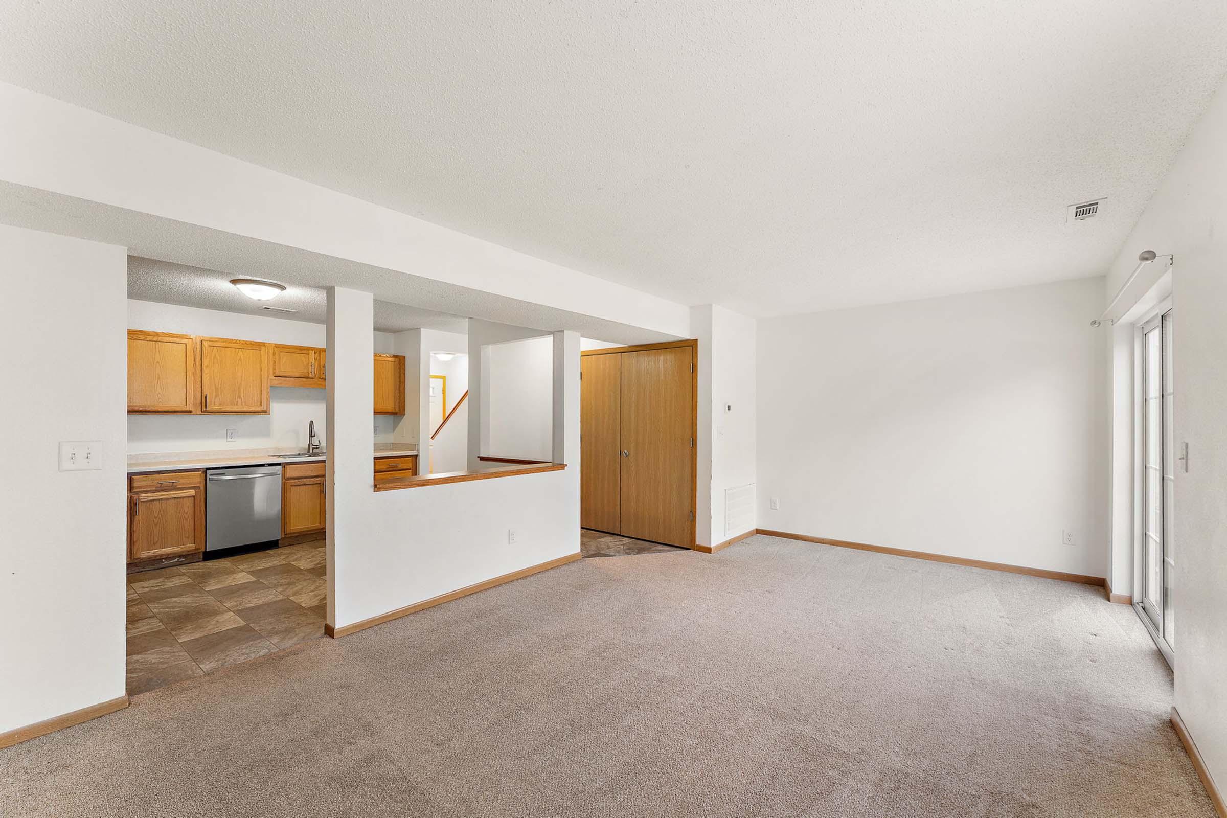 Interior view of a spacious living area featuring light-colored walls and carpet. A kitchen is visible with wooden cabinets and stainless steel appliances. There is a doorway leading to another room, and large sliding glass doors provide natural light. The layout is open and airy.