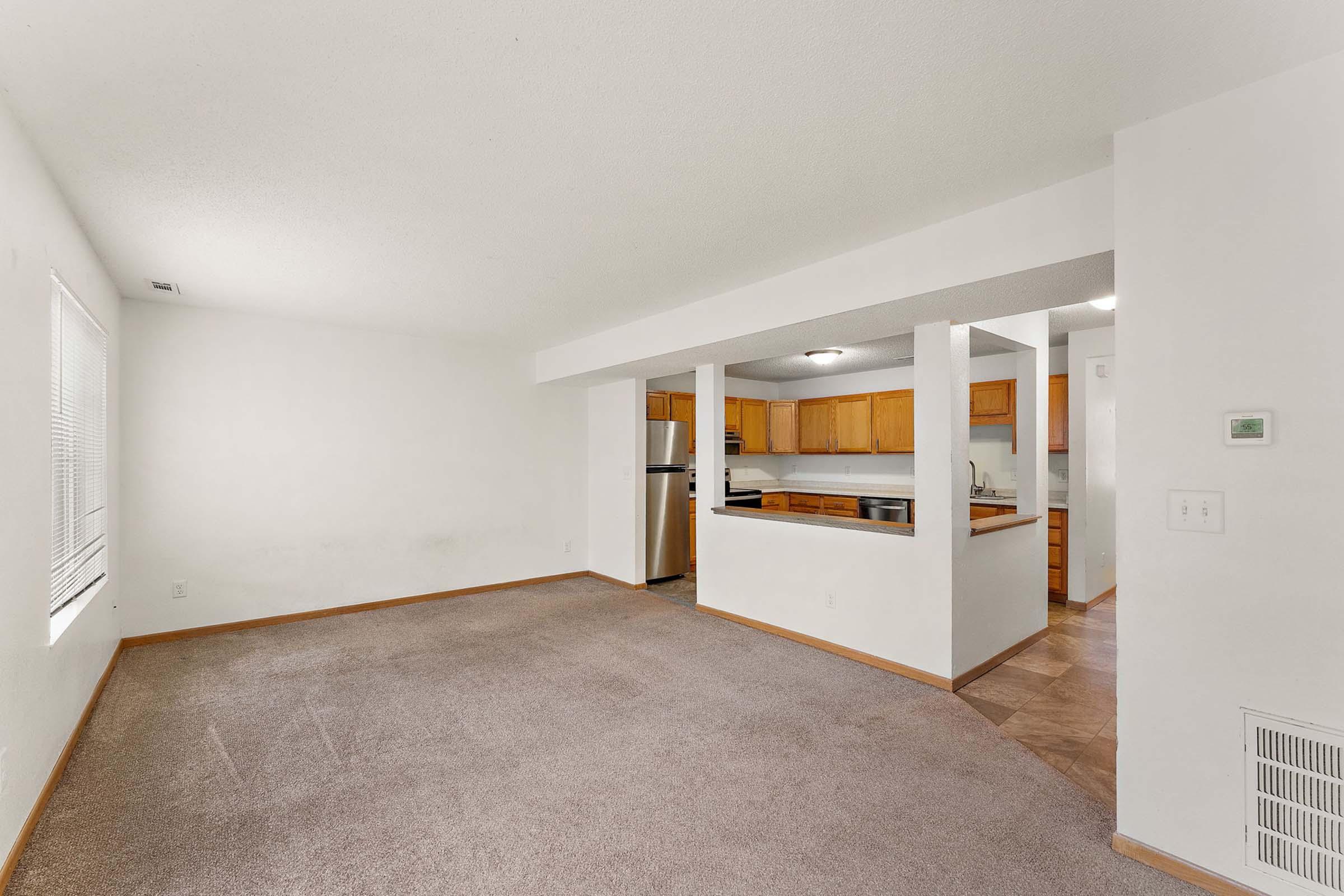 A spacious, empty living room with light-colored walls and a carpeted floor, featuring an open layout that connects to a kitchenette with wooden cabinets. Natural light comes through a nearby window, illuminating the area. The kitchen is partially visible in the background, showcasing appliances and counter space.