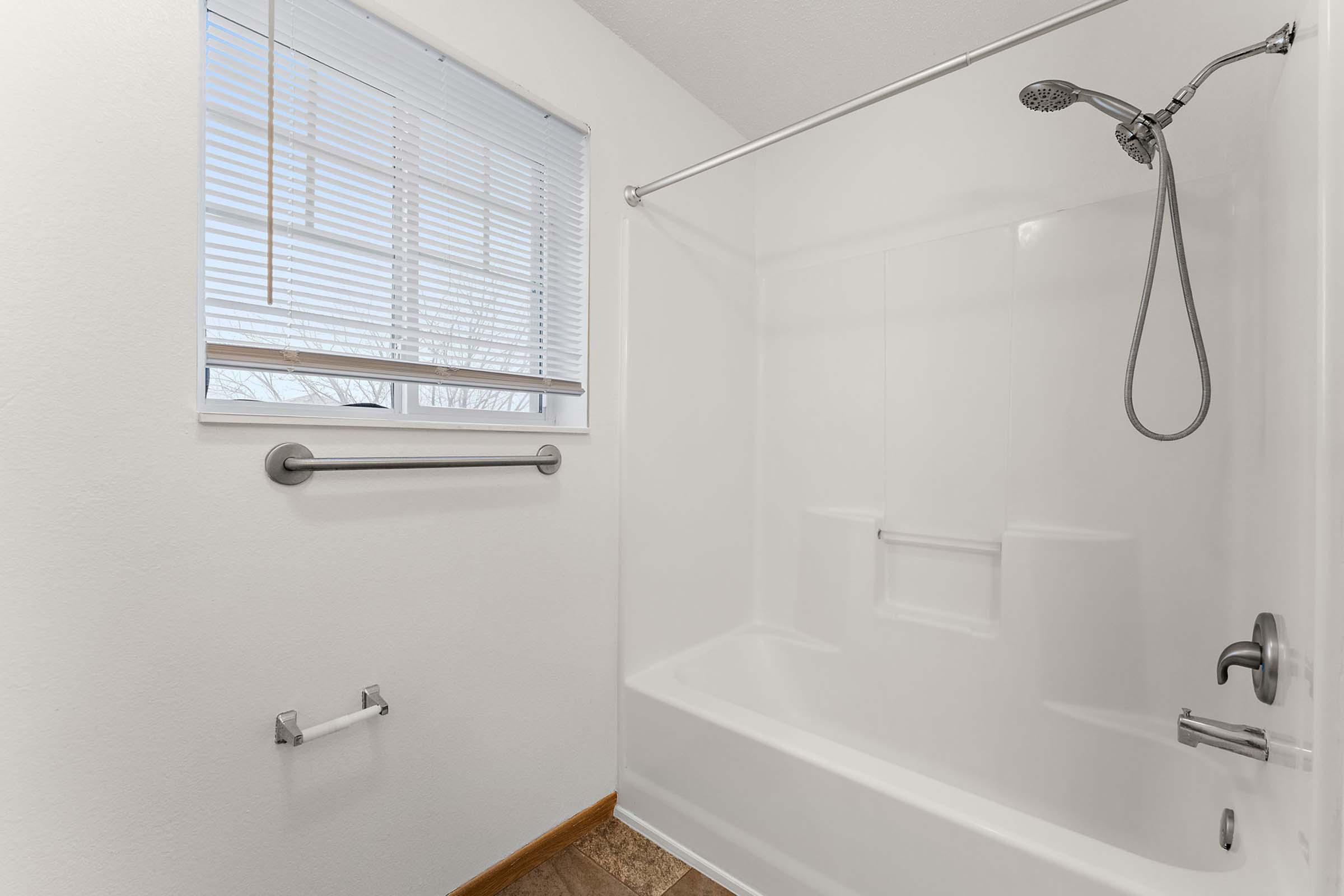 A clean, minimalist bathroom features a white shower tub combo, a chrome showerhead, and a window with blinds. There is a grab bar beside the tub and a toilet paper holder on the wall. The walls are painted white, and the floor is a light tan tile, creating a bright and airy atmosphere.