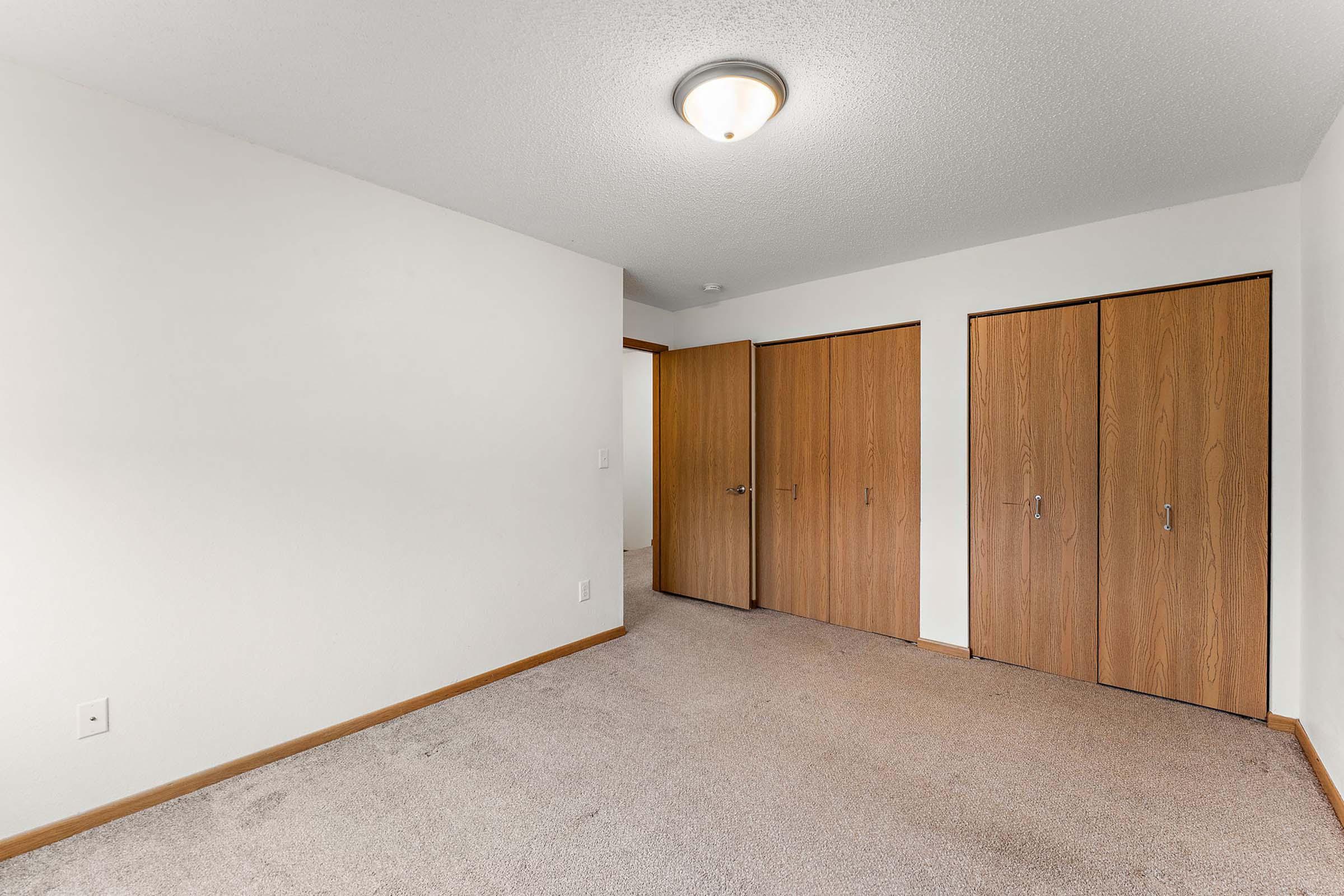 Empty room with beige carpet, white walls, and a ceiling light. Two wooden doors are visible, leading to closets, and the room has a neutral color scheme, providing a spacious and inviting feel.
