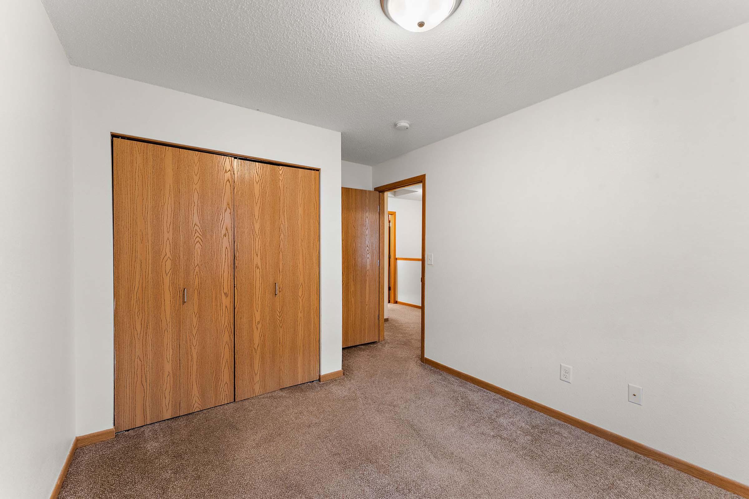 Interior view of a room featuring beige walls and carpet flooring. On the left, there are two wooden closet doors. In the background, a door leads to another room, with soft lighting from an overhead fixture. The overall setting appears clean and well-maintained.