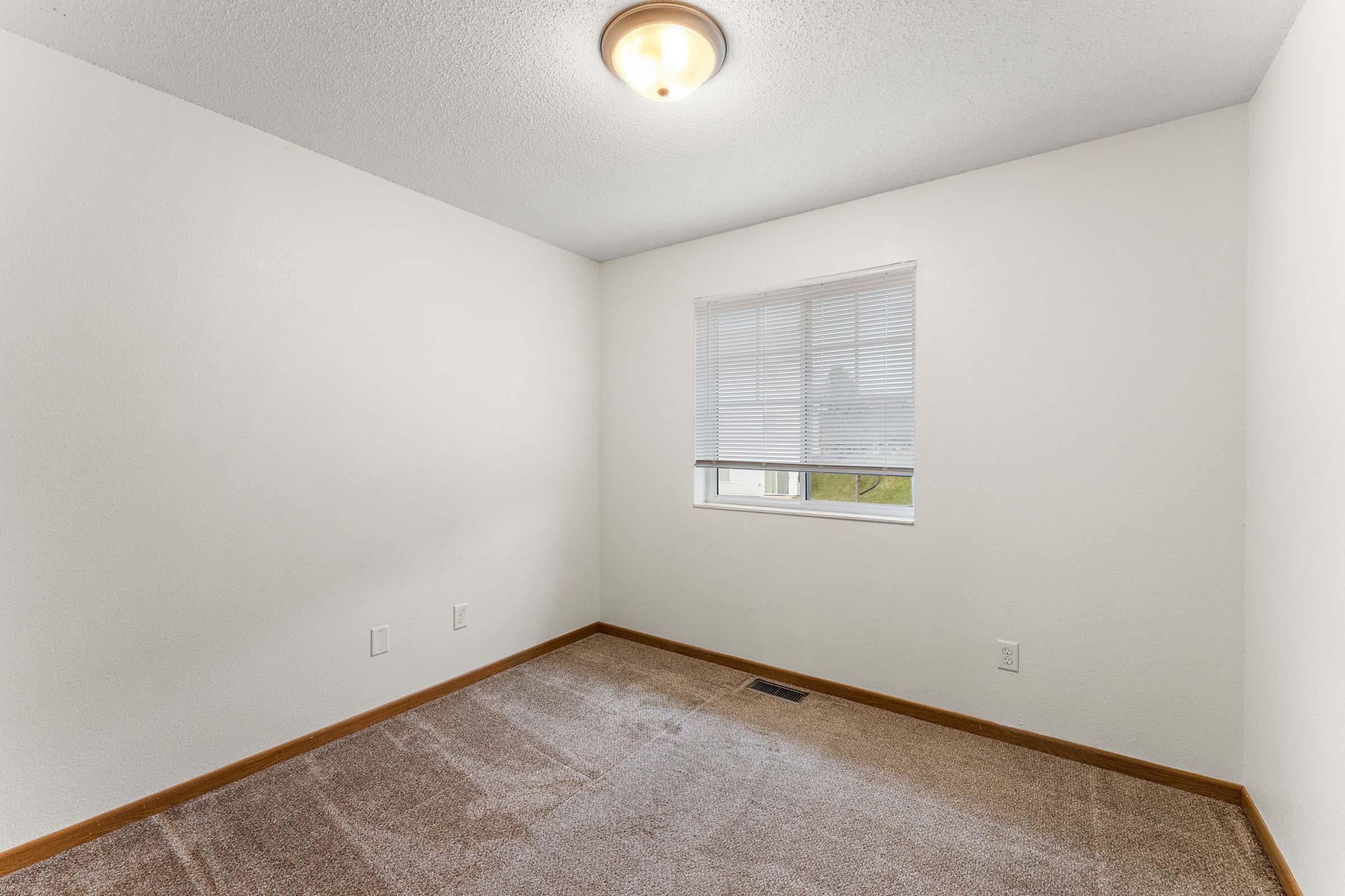 A small, empty room with beige carpet and white walls. A single window covered by blinds allows natural light to enter. The ceiling features a round light fixture. There are two small electrical outlets on one wall, and the overall feel of the room is bright and minimalistic.