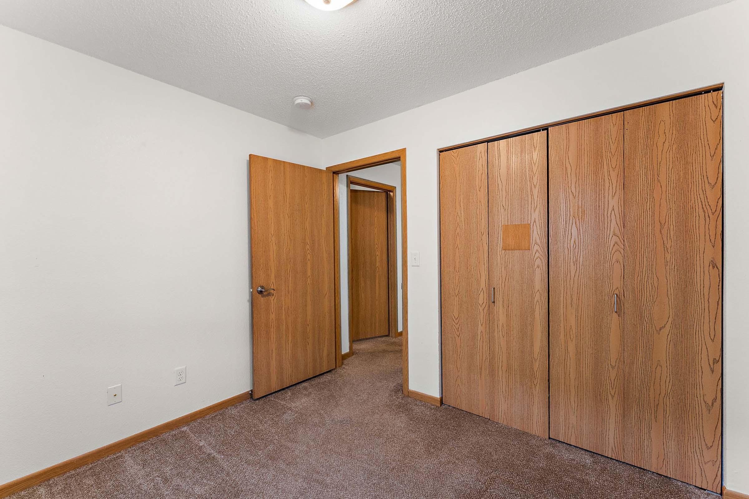 Empty room featuring a light-colored wall and brown carpet. A doorway leads to another room, and there are wooden closet doors on one side. The overall atmosphere is neutral and inviting, with good lighting from a ceiling fixture.