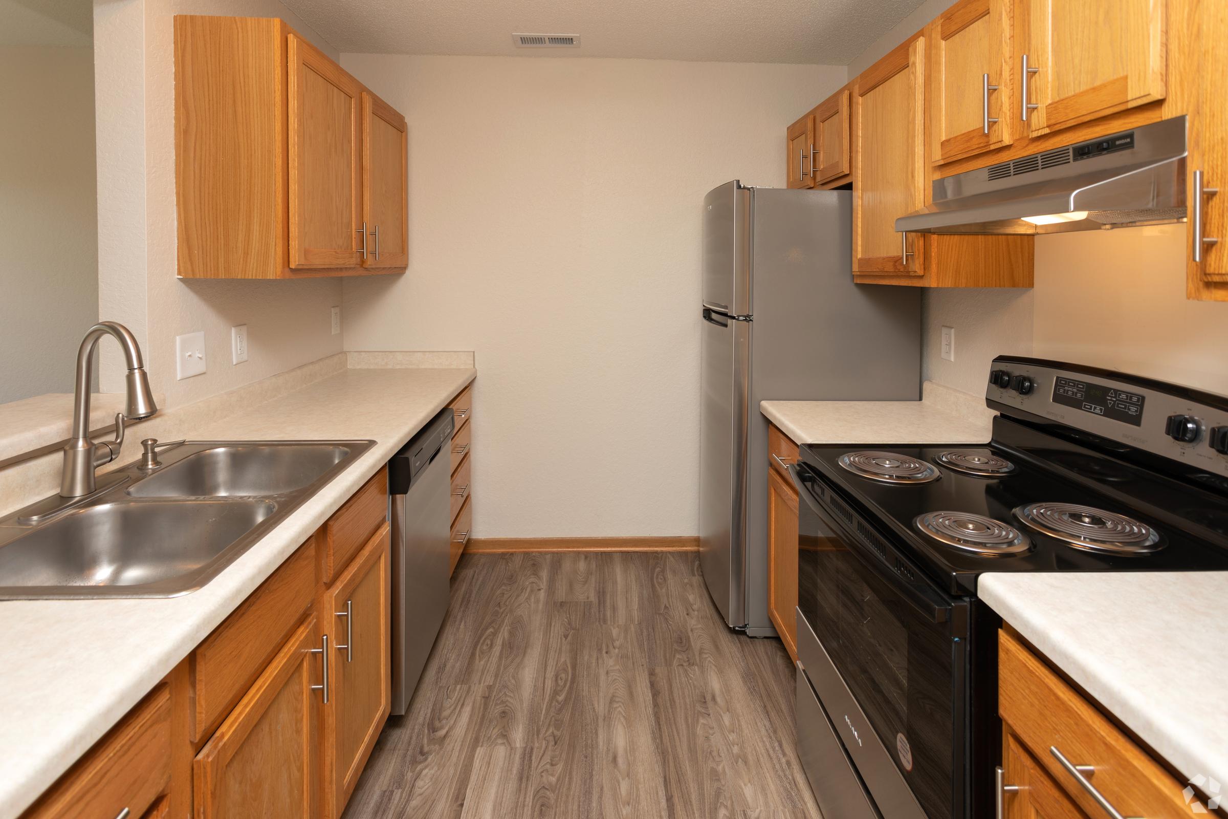 A kitchen featuring wooden cabinets, a double sink, stainless steel appliances including a refrigerator and an oven, and a smooth countertop. The floor is a light-colored laminate, and the walls are painted in a neutral shade, creating a modern and clean atmosphere.