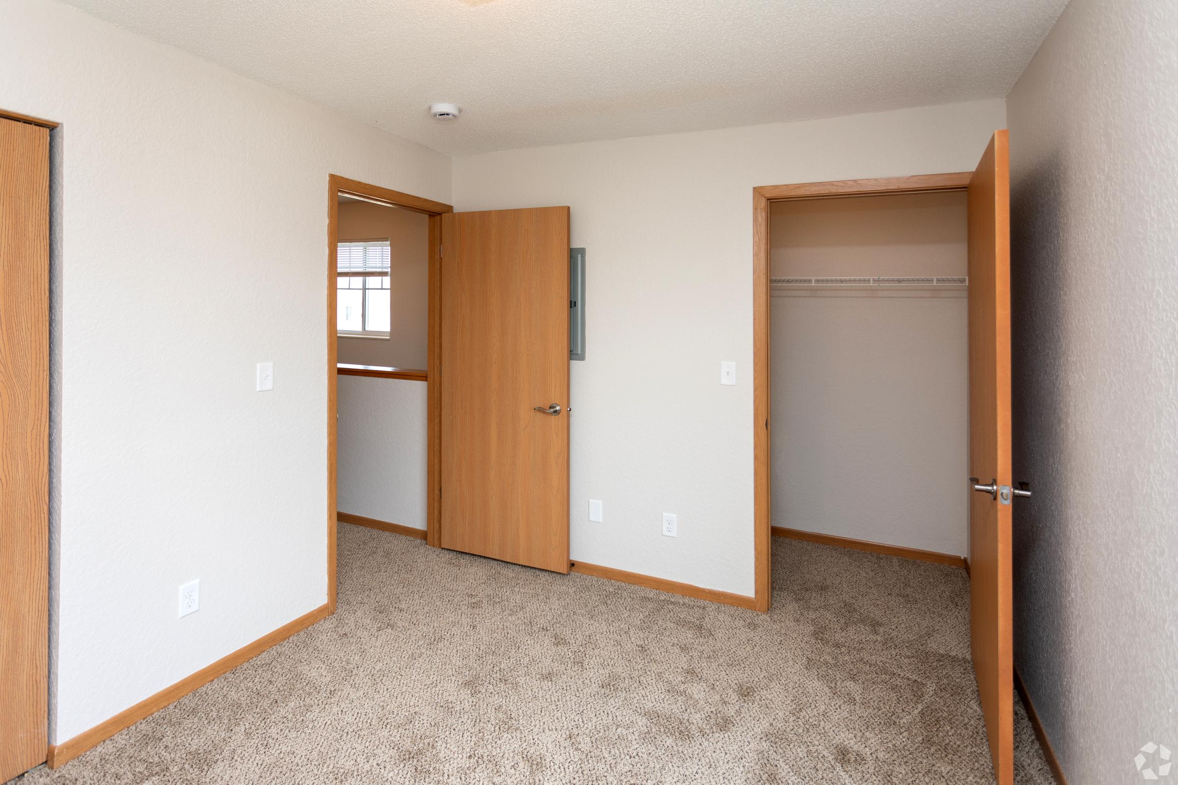 A spacious, empty bedroom featuring light-colored walls and soft carpeting. Two wooden doors lead to a closet and another room. Natural light streams through a window, highlighting the room's neutral tones and inviting atmosphere. The layout is simple, providing potential for various furniture arrangements.
