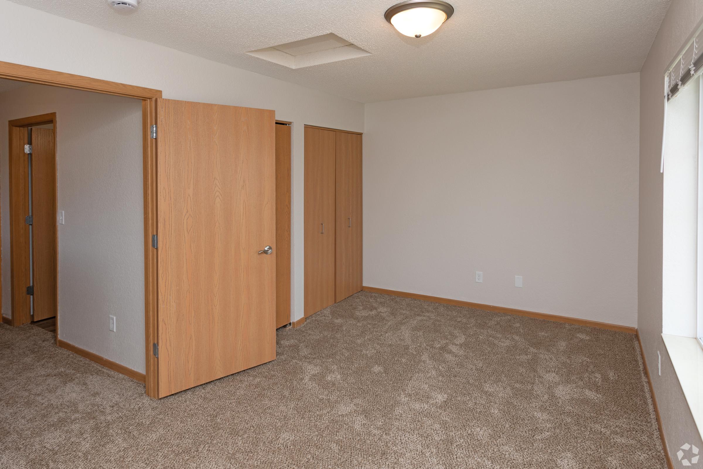 Empty room with beige walls and light brown carpet. Two wooden doors are visible, one leading to a closet and another to another room. A window is present on one wall, and there is a ceiling light fixture. The overall atmosphere is clean and modern, suitable for various uses.