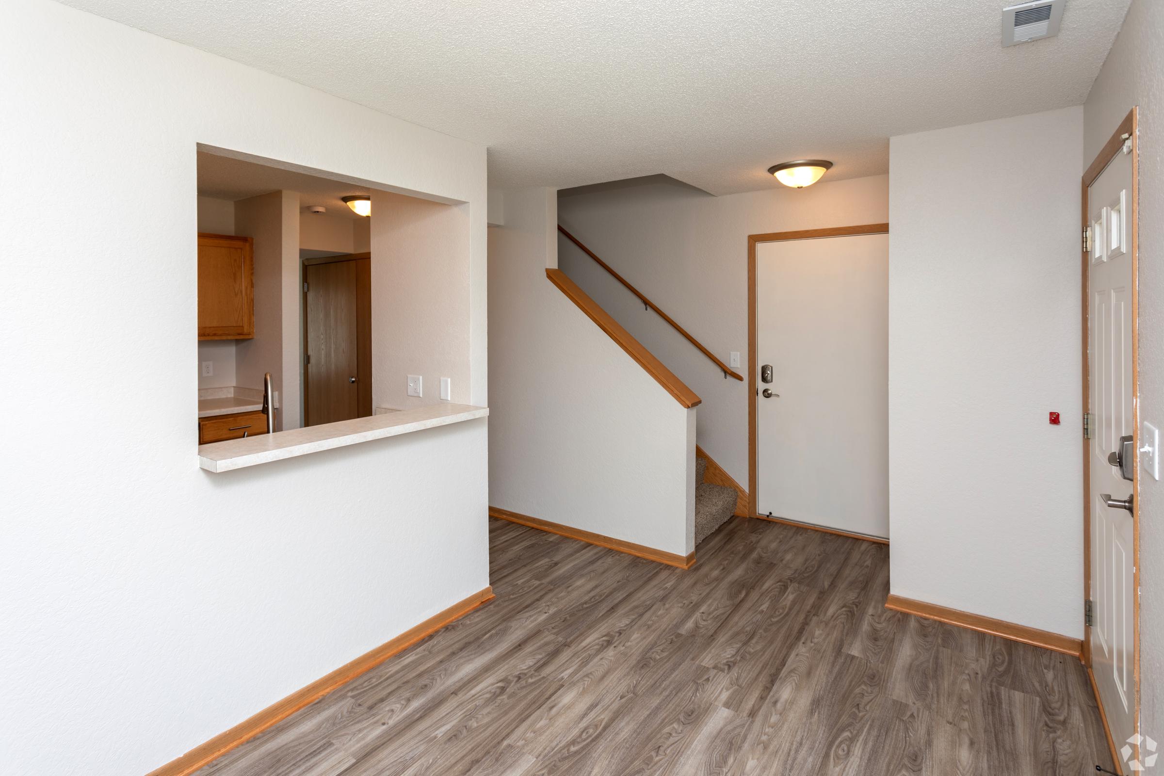 A modern entryway with light-colored walls and hardwood-style flooring. There's a stairway on the right leading to the upper level, and an open kitchen area visible through a pass-through opening. The door to the outside is located to the right. Overall, the space is bright and inviting.