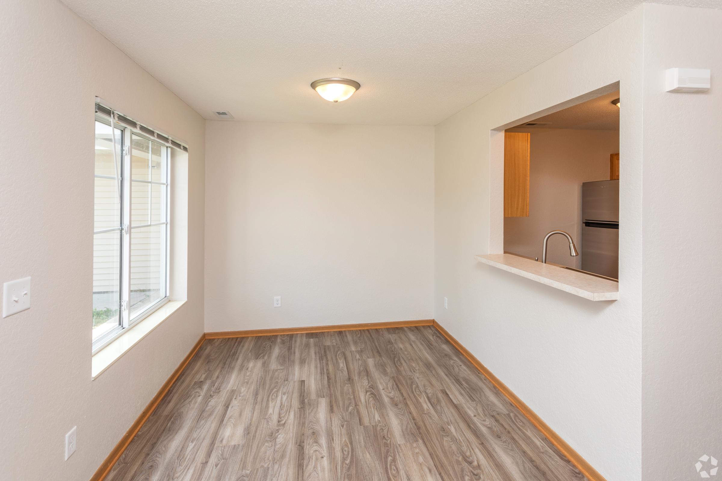 A small, empty room with light-colored walls and wooden trim. There's a large window on one wall, allowing natural light to enter. An opening leads to a kitchen area, featuring a light-colored counter and a glimpse of appliances. The flooring is a wood-like laminate.