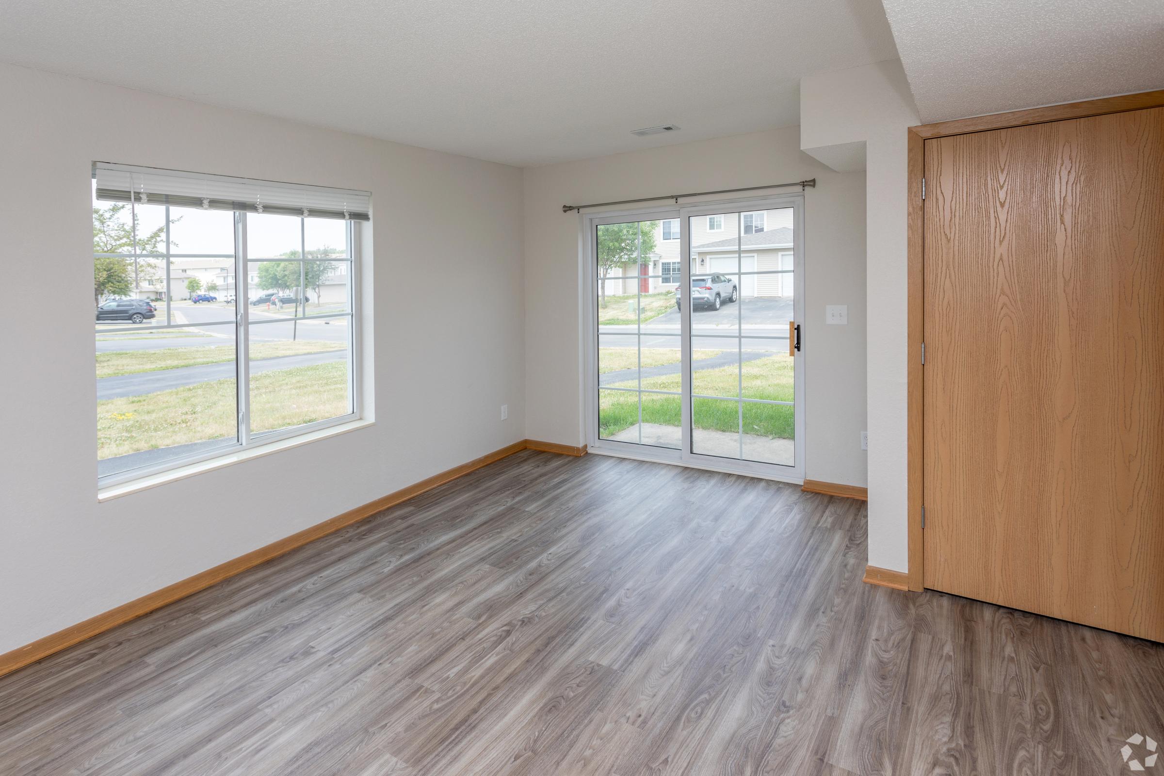 Interior view of a spacious, empty living room featuring light-colored walls, a large window letting in natural light, and a sliding glass door leading to an outdoor area. The floor is covered with modern, gray vinyl flooring, and there is a small section of wood paneling.