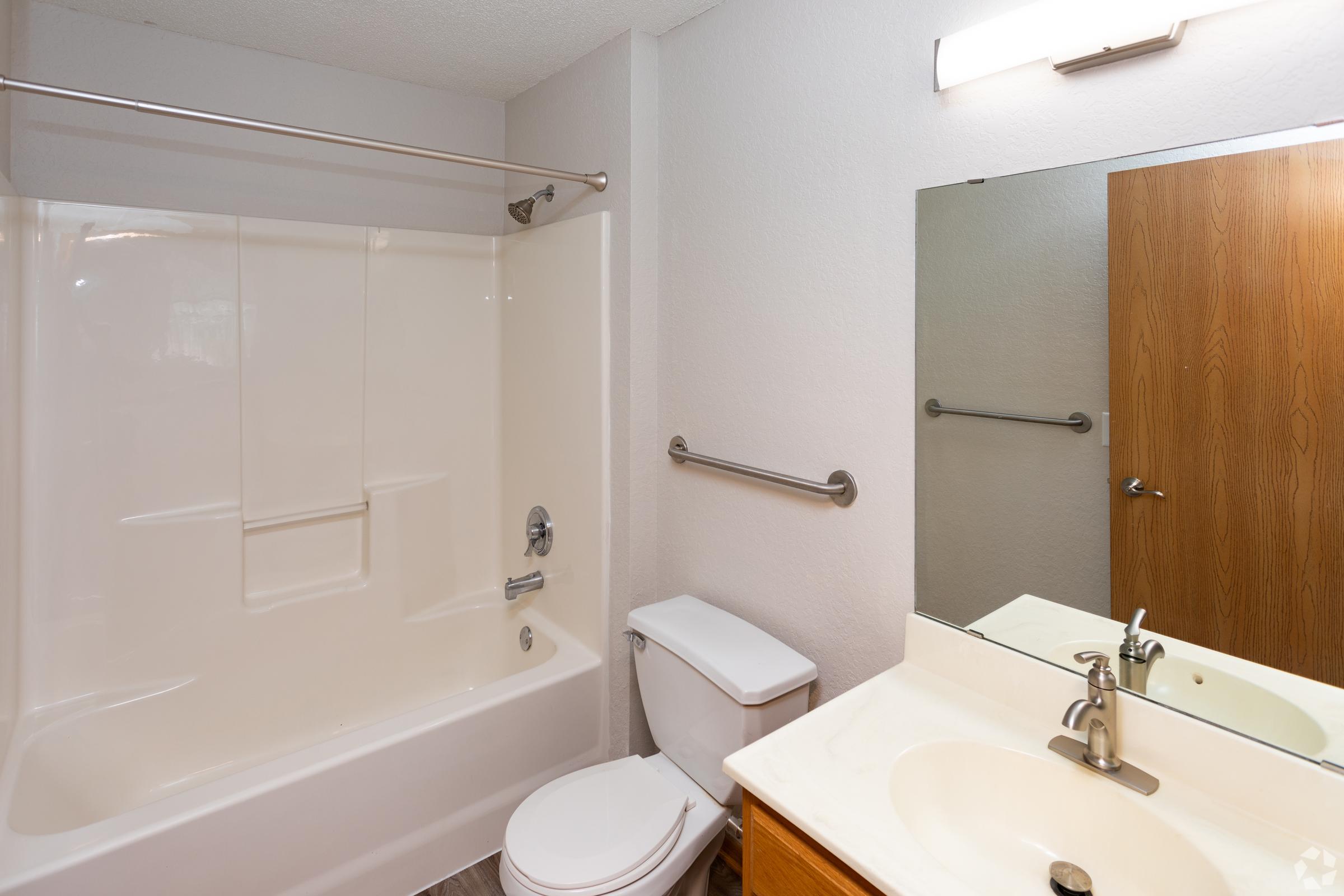 A clean and modern bathroom featuring a white bathtub with a shower, a toilet, and a sink with a countertop. The walls are painted in light colors, and there's a mirror above the sink. A wooden cabinet is visible, and there's good lighting from an overhead fixture.