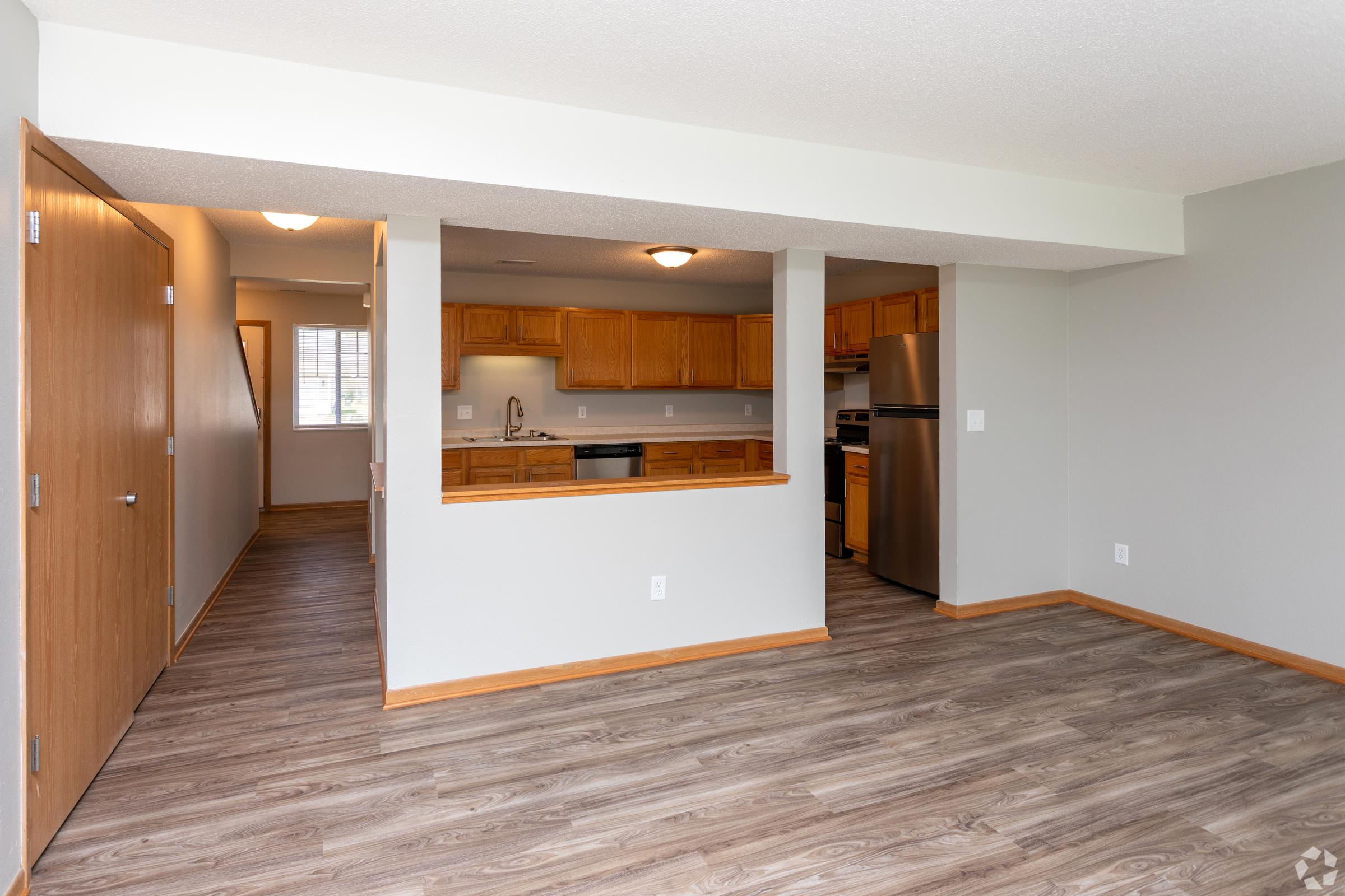 Spacious living area with beige walls and laminate flooring, featuring an open layout that connects to a kitchen with wooden cabinets and stainless steel appliances. Natural light enters through a window in the background, enhancing the bright and inviting atmosphere.