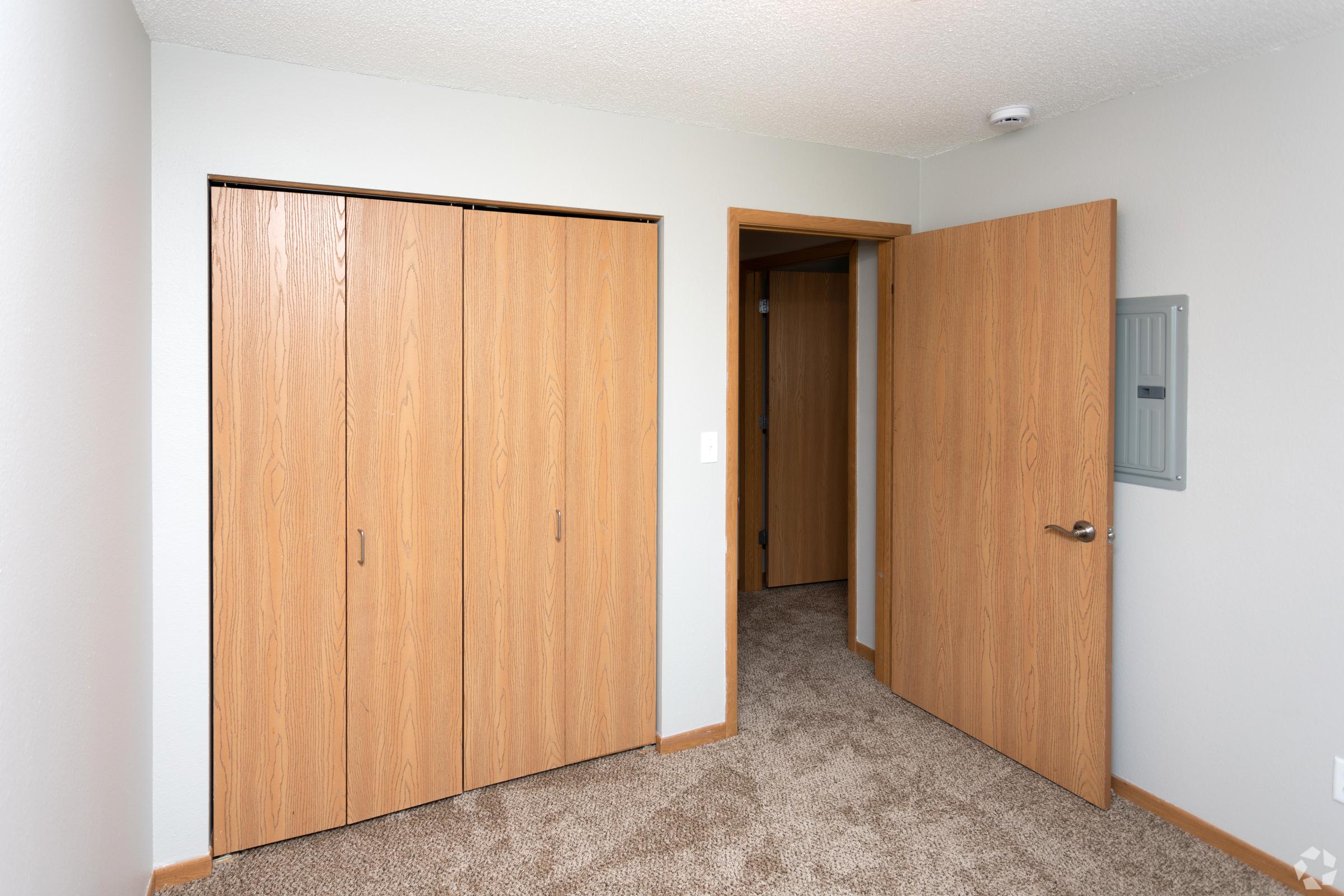 A well-lit interior space featuring a beige carpeted floor. On the left, a set of wooden sliding doors leads to a closet, while on the right, an open door reveals a hallway. The walls are painted light gray, and there's a utility box mounted on the wall next to the door.