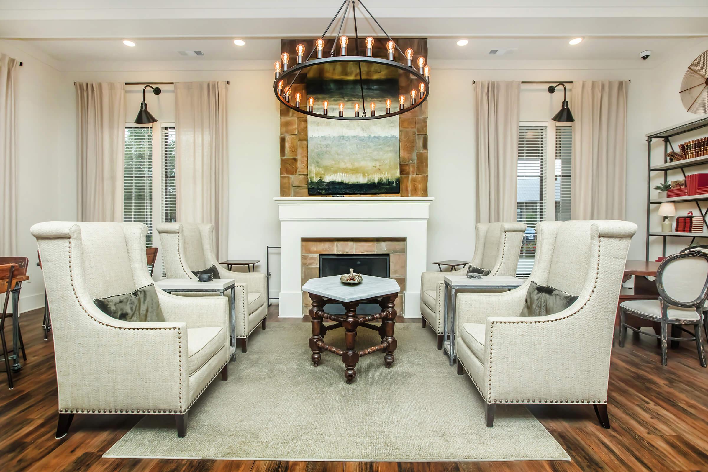 A modern living room features four upholstered armchairs arranged around a round coffee table on a plush area rug. There's a fireplace with a decorative mantel, and a large circular chandelier hangs above. Light-colored curtains frame the windows, and a large abstract painting is displayed above the fireplace.