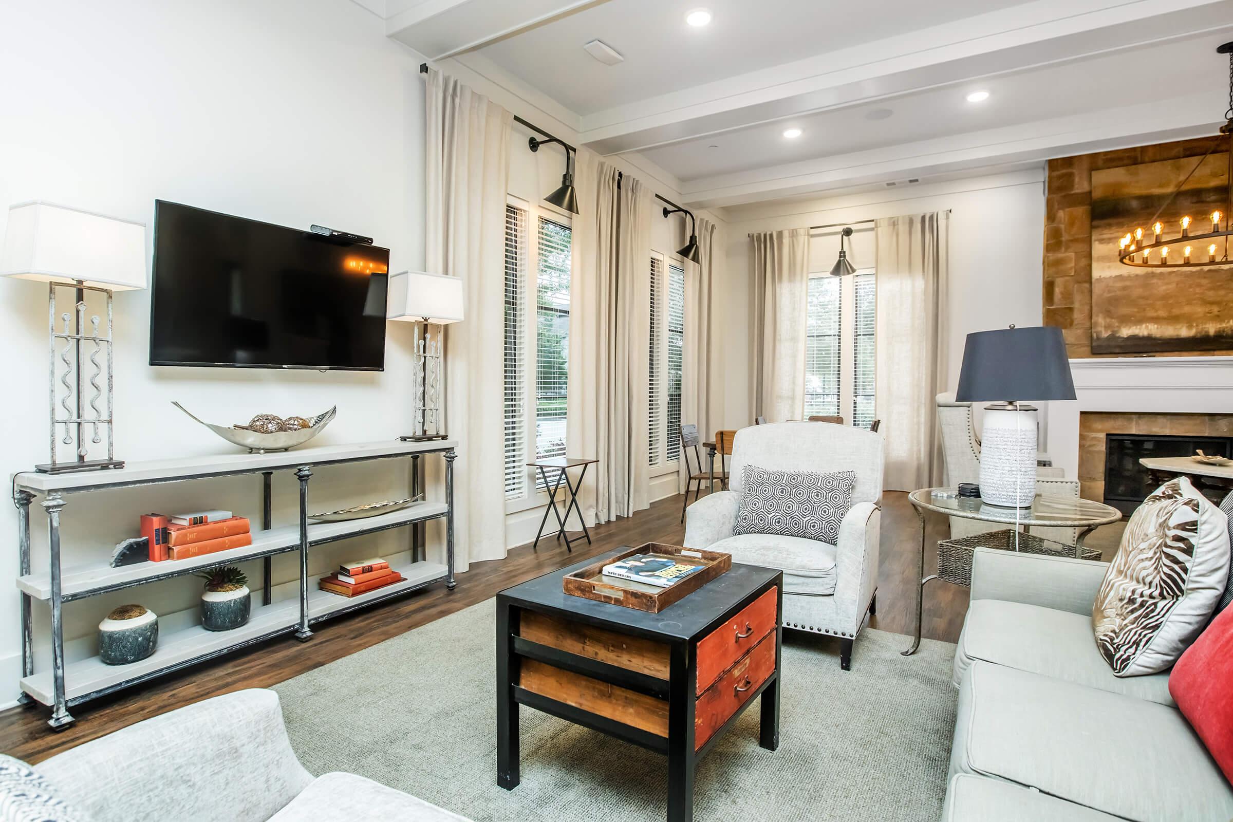 A cozy living room featuring a comfortable armchair and sofa, a wooden coffee table with books, a modern TV mounted on the wall, and decorative lamps. Large windows with sheer curtains allow natural light, and a stone fireplace adds warmth to the space. Stylish decor elements create an inviting atmosphere.