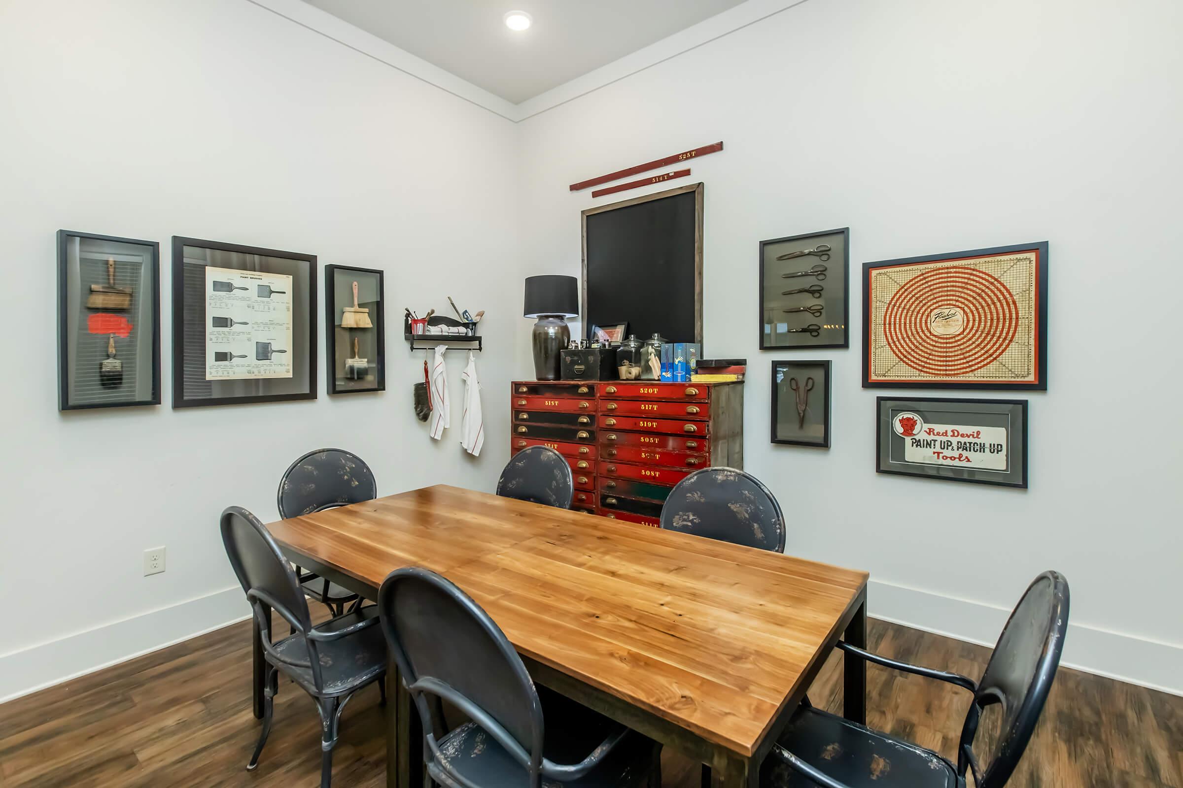A spacious room featuring a wooden table surrounded by black chairs. The walls are adorned with various framed artworks and tools, including sketches and a large circular target design. A red tool chest is positioned against one wall, enhancing the workshop-like atmosphere.