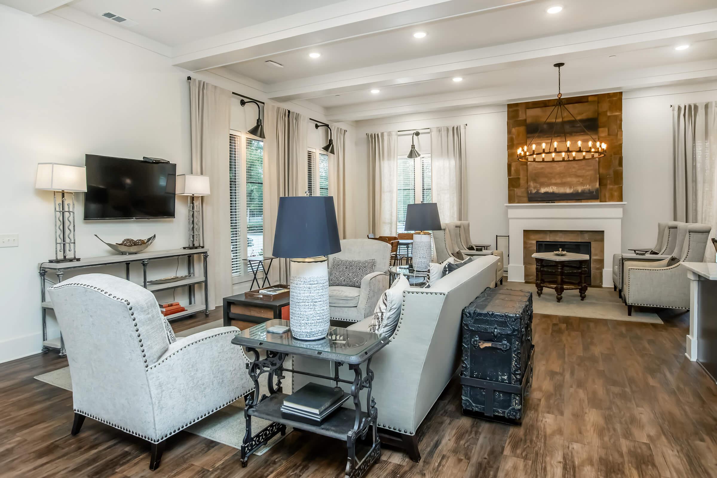 A modern living room featuring light-colored walls and a wood floor. It has stylish gray armchairs and a comfortable sofa, a coffee table, and decorative lamps. A TV is mounted on the wall, and there’s a decorative fireplace with a chandelier overhead, along with large windows adorned with sheer curtains.