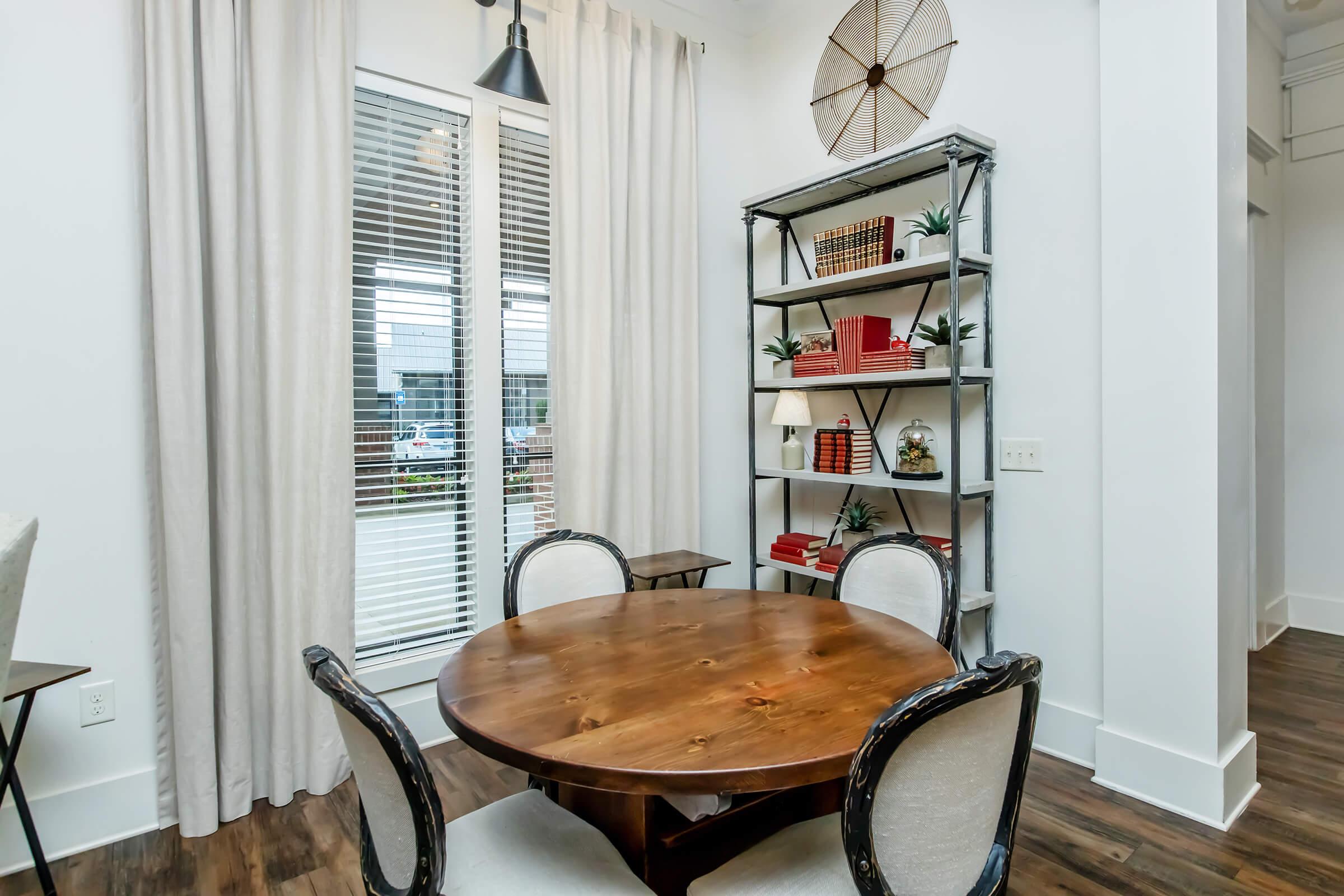 A cozy dining area featuring a round wooden table surrounded by four upholstered chairs. A tall bookshelf with decorative items and red books is adjacent to large windows with white curtains, allowing natural light to fill the space. The floor is dark wood, adding warmth to the room's modern aesthetic.