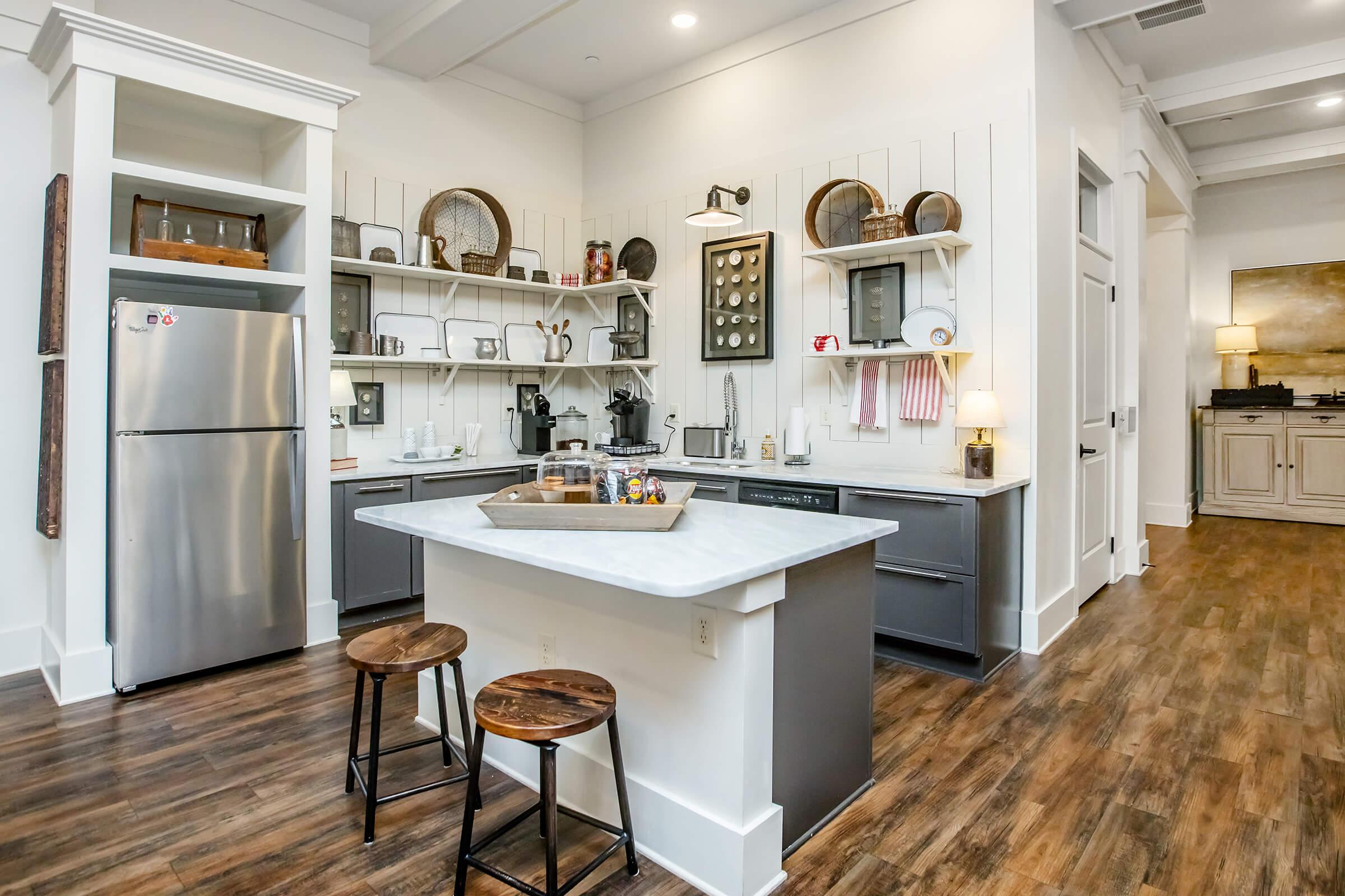 A modern kitchen with a marble-topped island, two wooden stools, stainless steel refrigerator, and open shelving displaying dishes and kitchenware. The decor features a bright, airy design with warm wood flooring and soft lighting, creating a welcoming atmosphere.