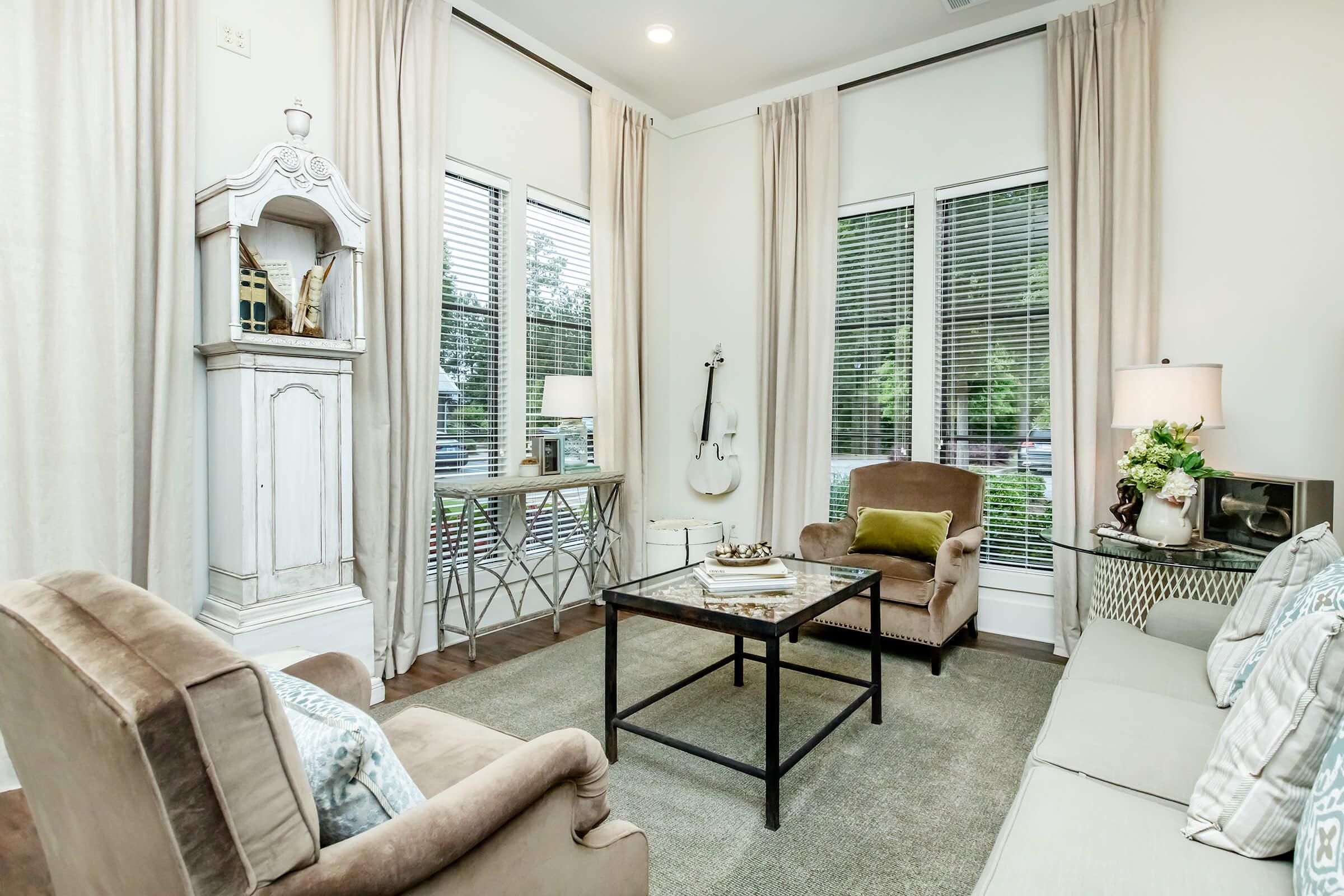 A cozy living room featuring two beige chairs, a light gray sofa, and a glass coffee table on a soft carpet. There’s a decorative cabinet, a violin, and large windows with white curtains, allowing natural light to fill the space. Stylish decor elements create a warm and inviting atmosphere.