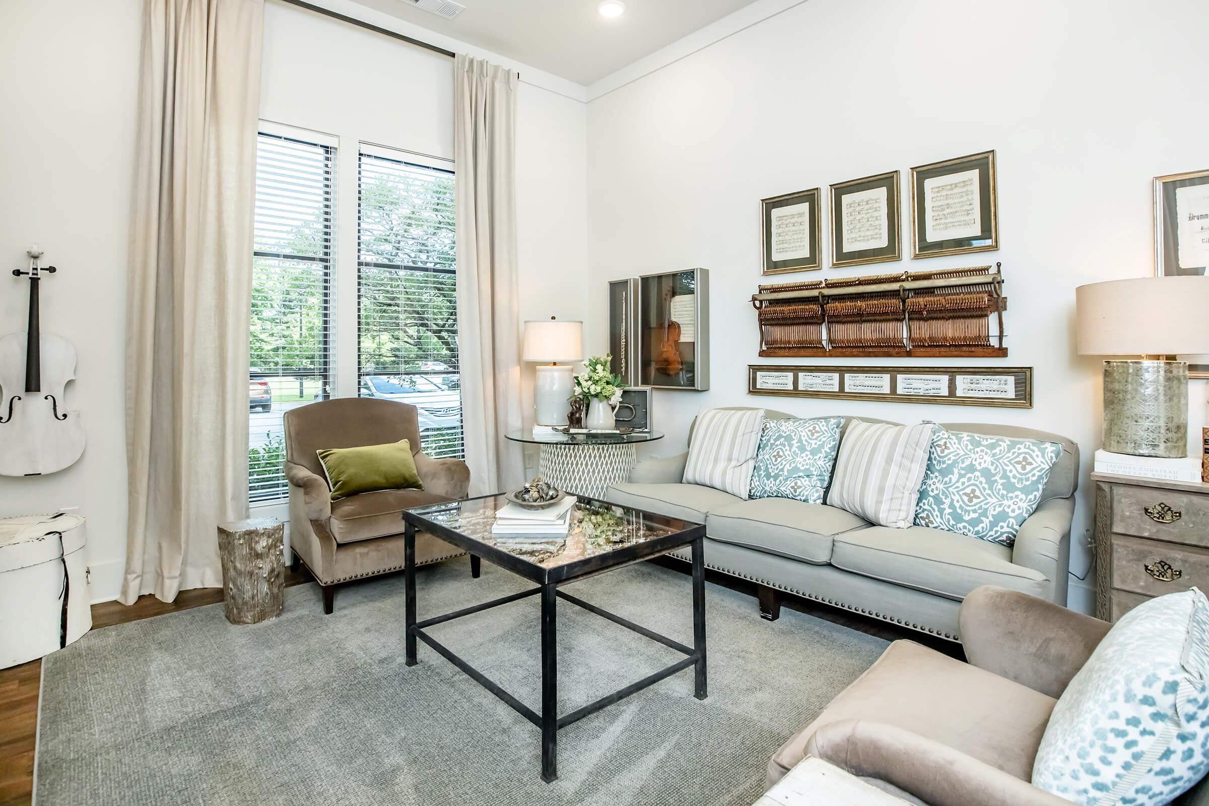 A cozy living room featuring a light-colored sofa with patterned cushions, two armchairs, a glass coffee table, and a decorative side table. The walls are adorned with framed artwork and a large window with curtains lets in natural light, creating a welcoming atmosphere.