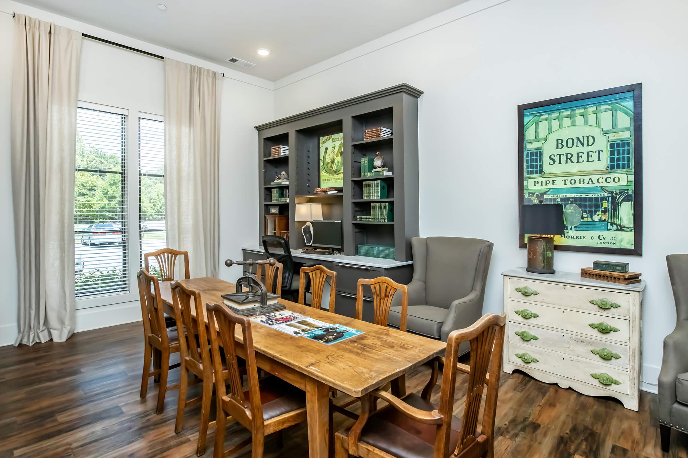 A cozy interior featuring a wooden dining table surrounded by eight chairs. There is a stylish bookshelf with decorative items and a vintage advertisement on the wall. Large windows allow natural light to fill the space, complemented by neutral-colored curtains and warm wooden flooring.
