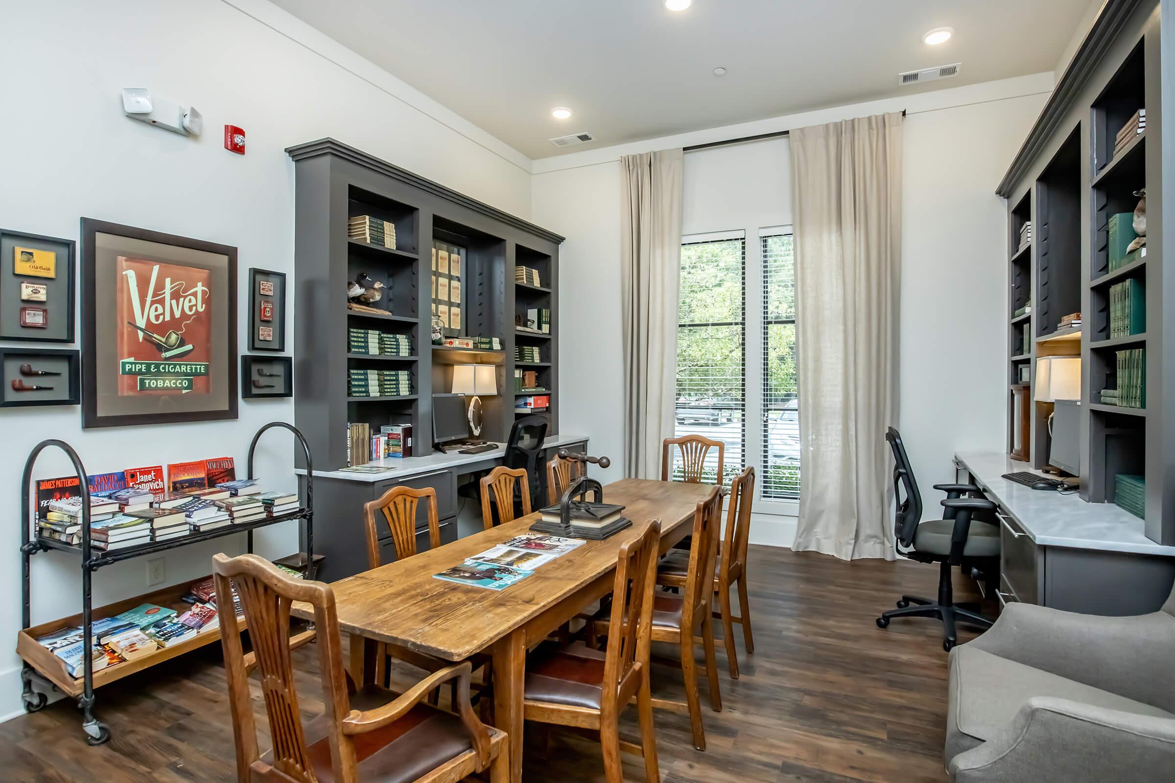 A stylish office space featuring a large wooden table surrounded by chairs, a gray couch in the corner, and built-in shelves filled with books and decorative items. Natural light comes through large windows with sheer curtains, creating an inviting atmosphere for work or meetings.