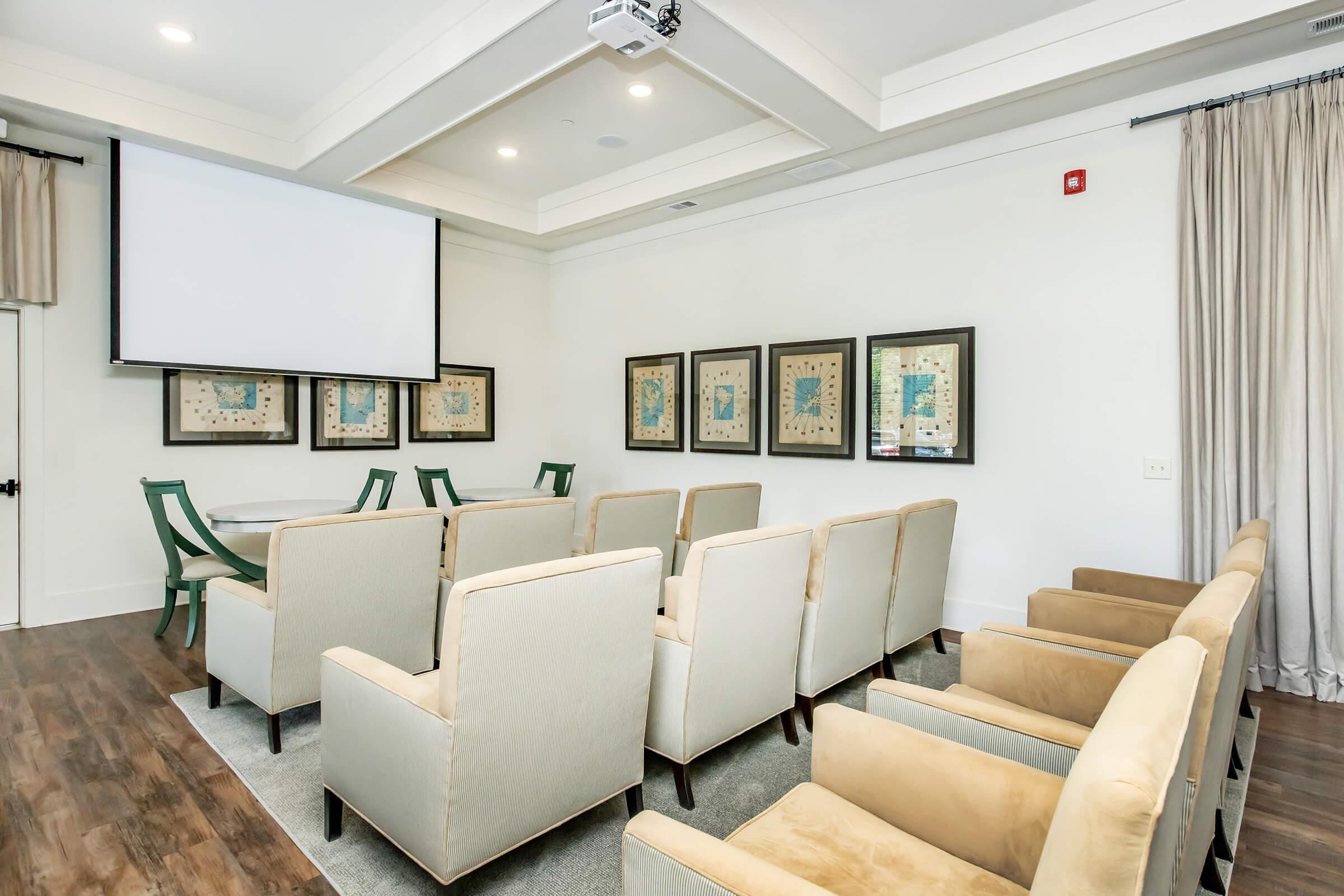 A modern, well-lit conference room featuring a projector screen at the front, surrounded by several framed artworks. There are rows of beige armchairs arranged in front of a round table, with green chairs visible in the background. The floor is wooden, and large curtains are drawn at the windows.