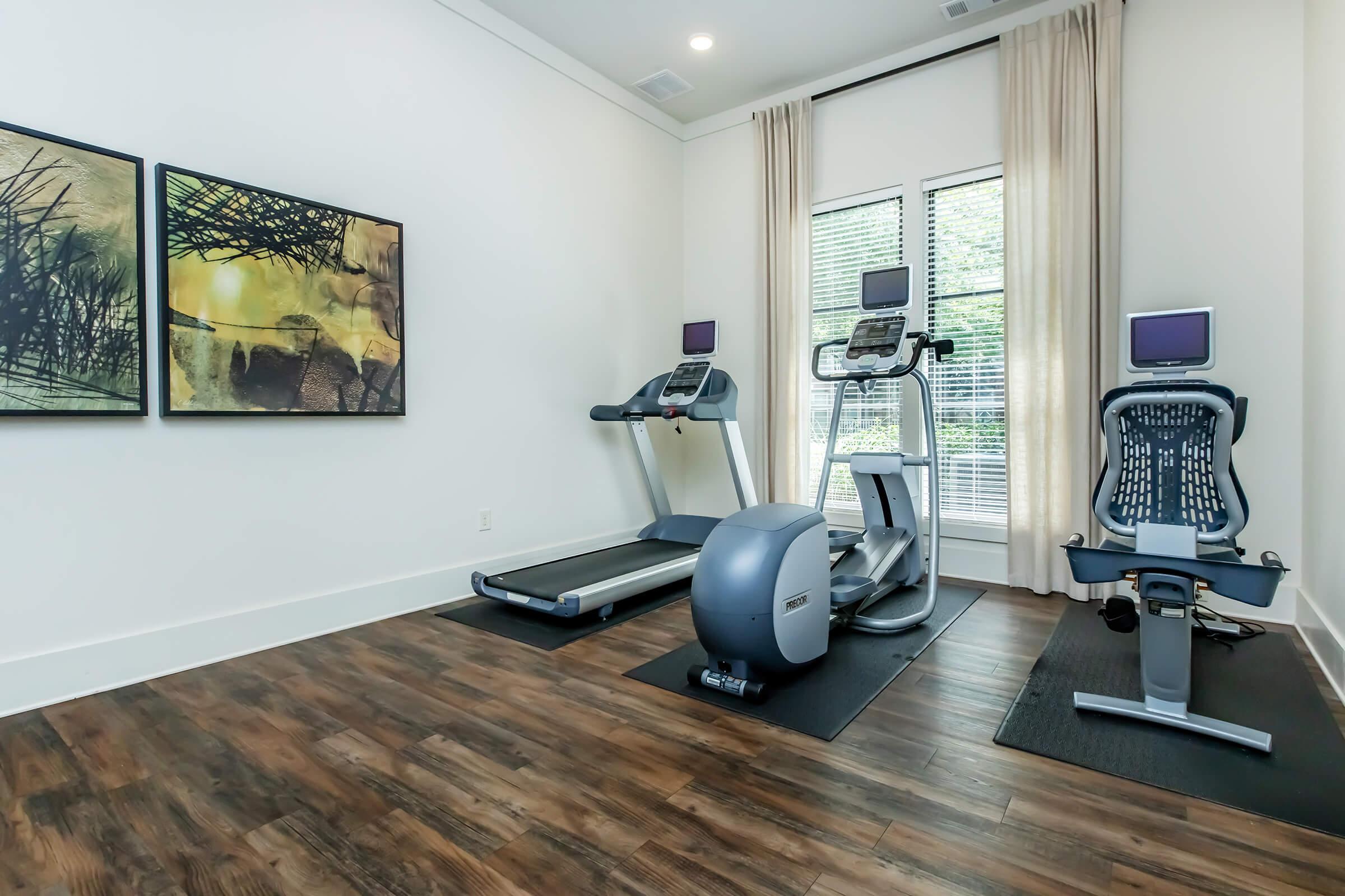 A modern home gym featuring two treadmills, a stationary bike, and an elliptical machine. The gym has large windows with natural light, and artwork on the walls. The floor is wooden, creating a warm and inviting atmosphere, with workout mats under the exercise equipment.
