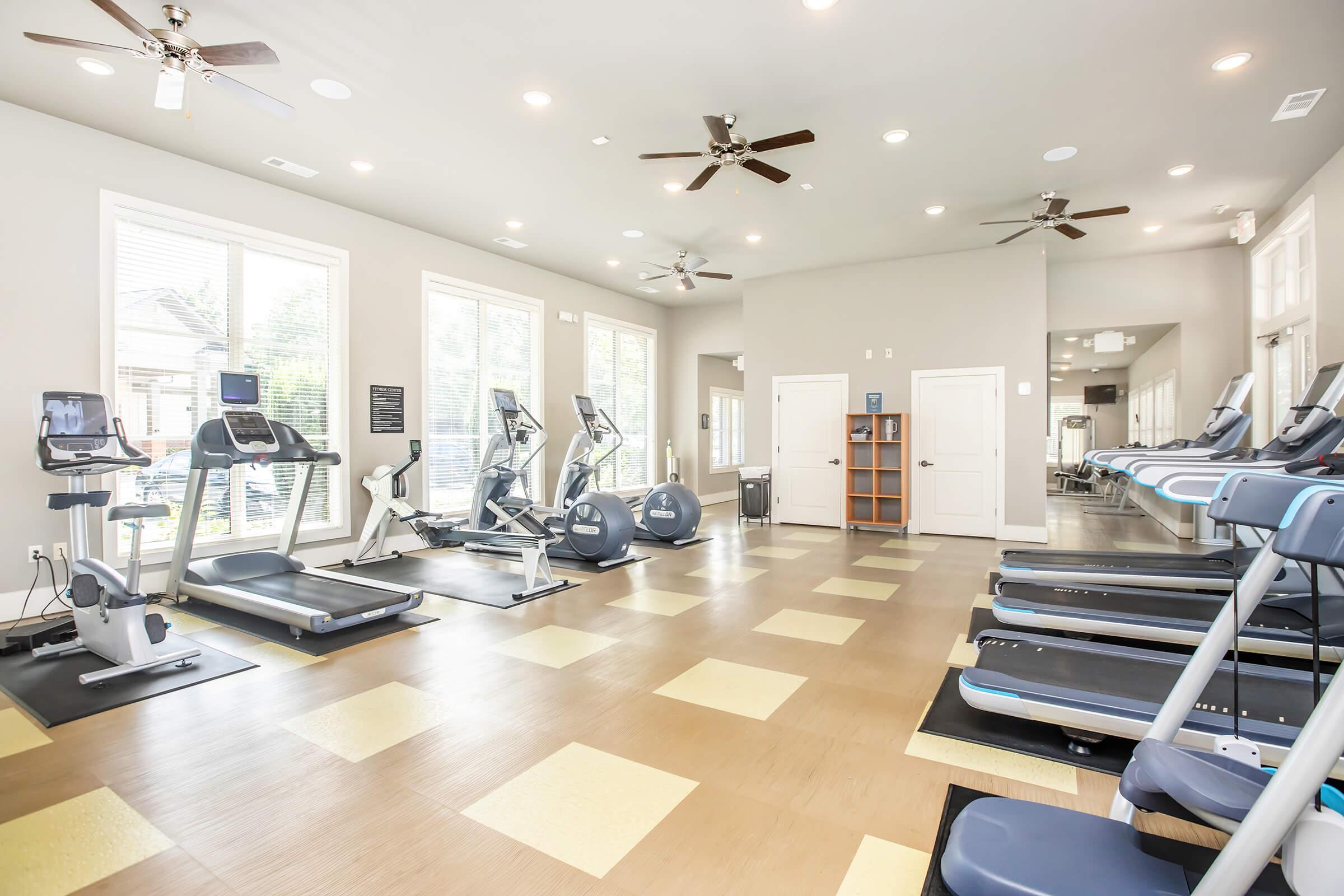 A spacious fitness center featuring several exercise machines, including treadmills and ellipticals, under bright lighting. Large windows provide natural light, and ceiling fans circulate air. The flooring is a mix of tiles, and the walls are painted in neutral colors, creating an inviting workout environment.